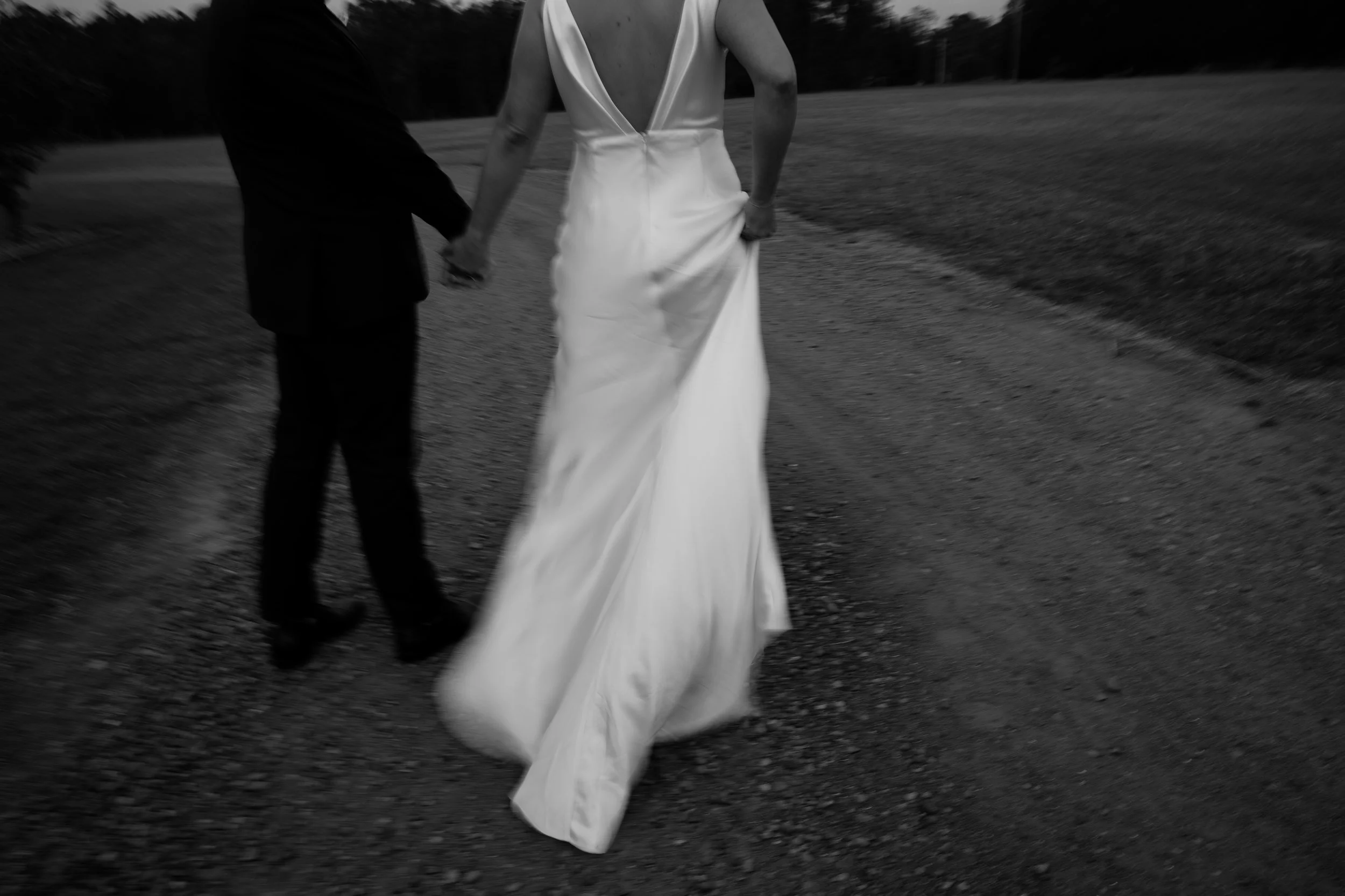 A person in a white wedding dress walking outdoors, holding hands with another person, on a dirt path with trees in the background.