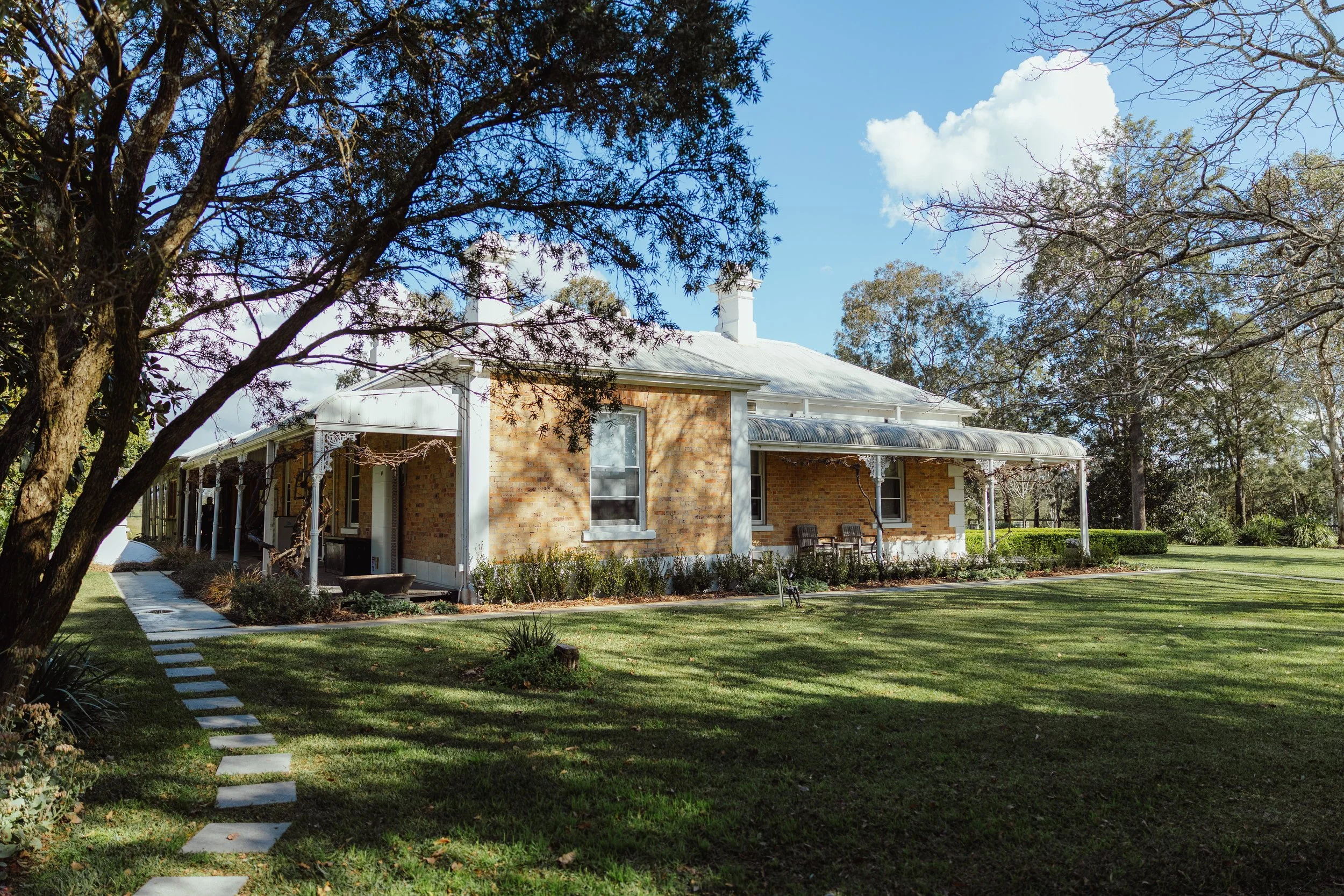 A brick house with a white metal roof and a front porch, situated in a lush green yard with trees and a walkway.