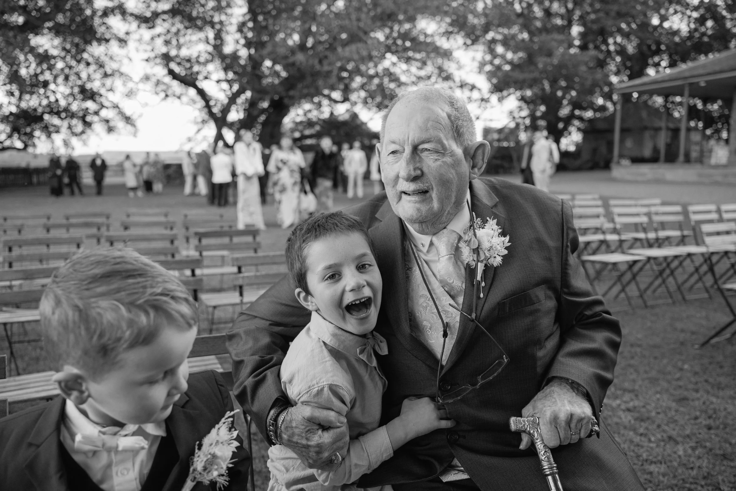 An elderly man in a suit with a boutonniere and a young boy smiling and sitting close together at an outdoor event, with other people and benches in the background.