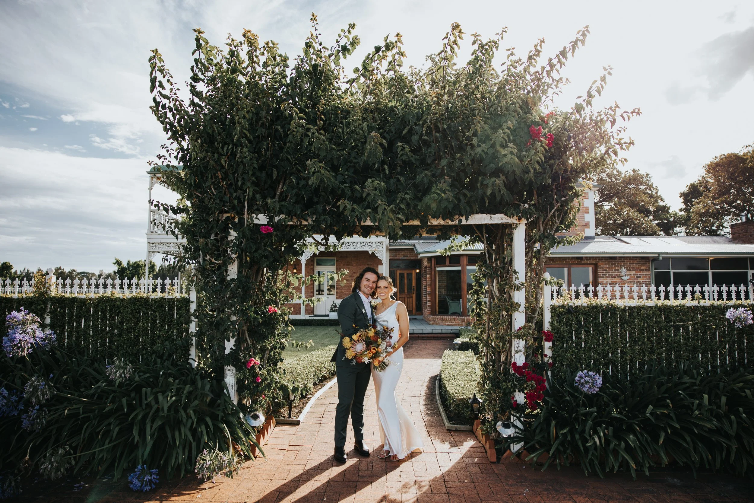 A newlywed couple standing under a garden arch made of greenery and flowers, smiling at the camera. The bride is in a white wedding dress holding a bouquet, and the groom is in a dark suit. The background includes a brick house with a porch, trees, a
