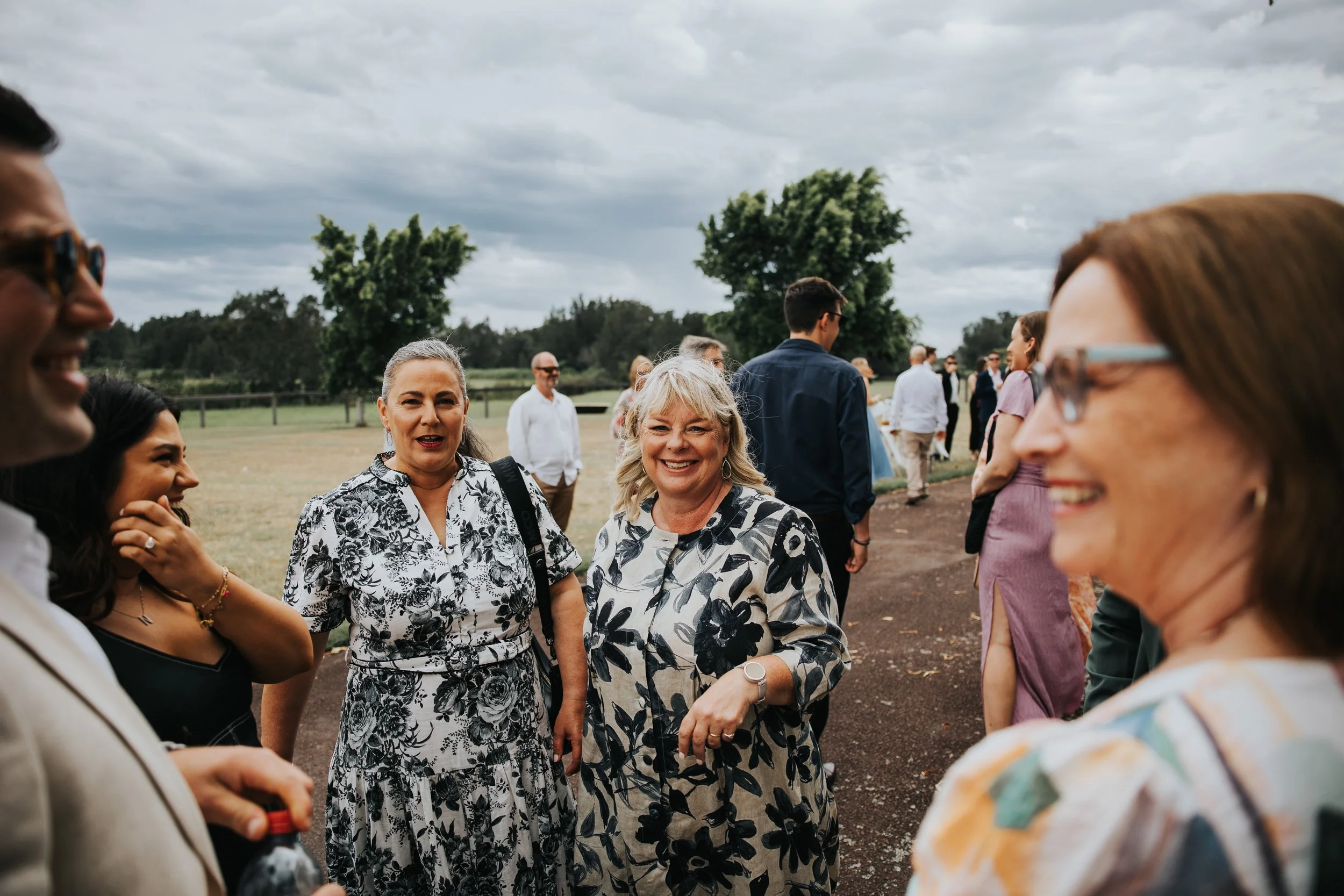 Group of people gathering outdoors on a cloudy day, smiling and talking, with some wearing sunglasses and casual clothes, standing on a dirt path with trees in the background.