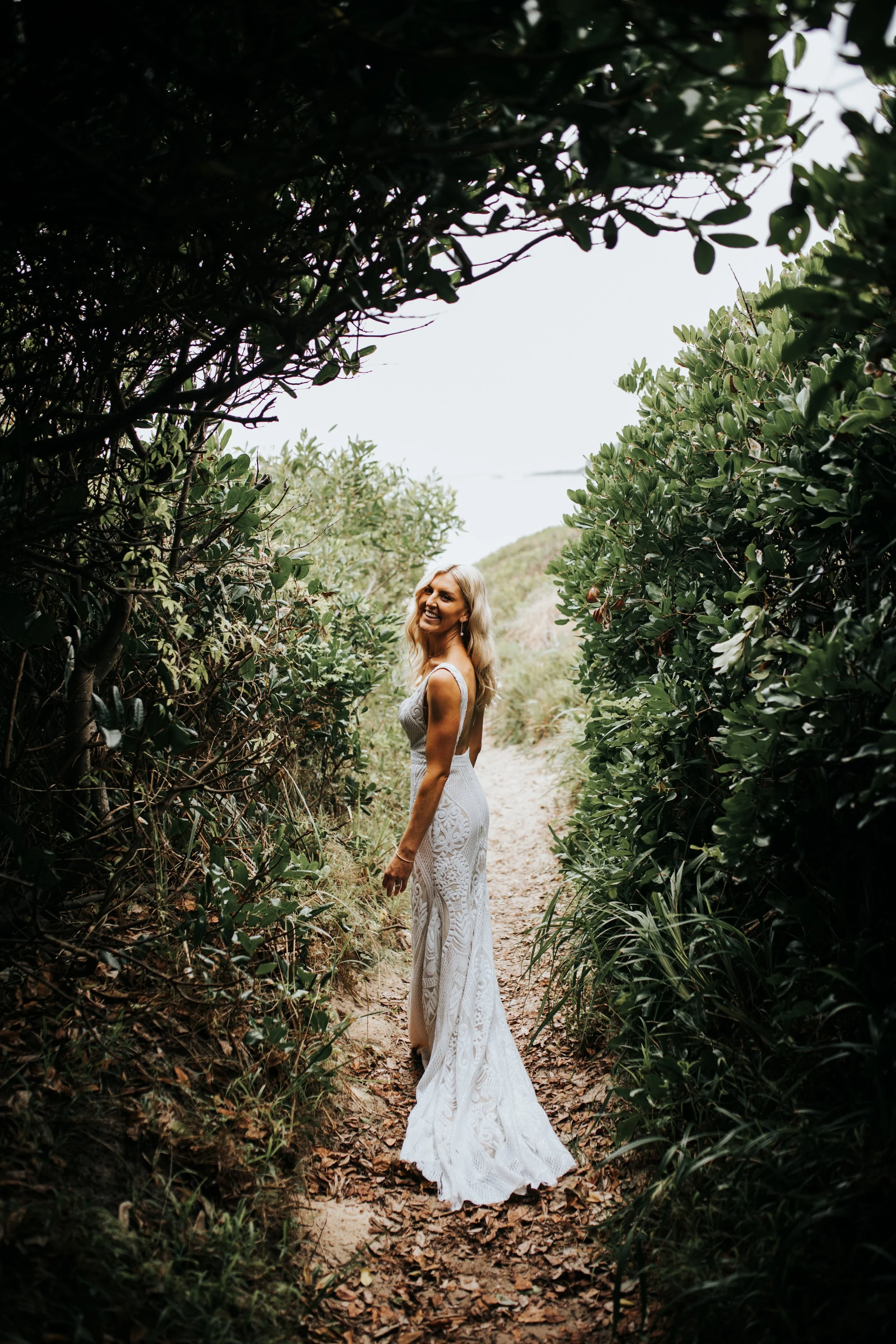 A woman in a white lace wedding dress standing on a sandy trail surrounded by dense green bushes and trees, smiling and looking back over her shoulder.