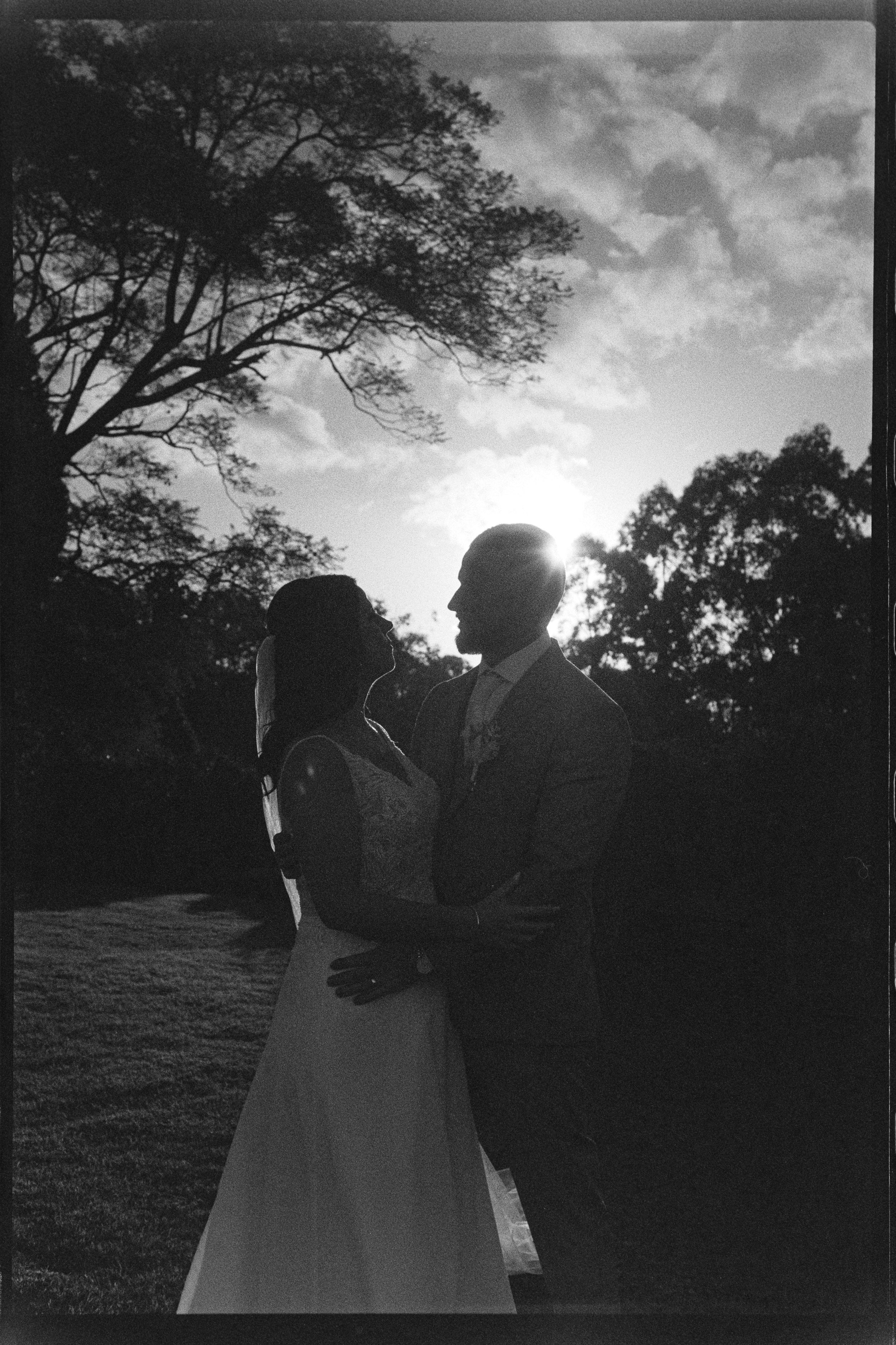 Silhouette of a bride and groom in wedding attire facing each other outdoors at sunset, with trees and a partly cloudy sky in the background.