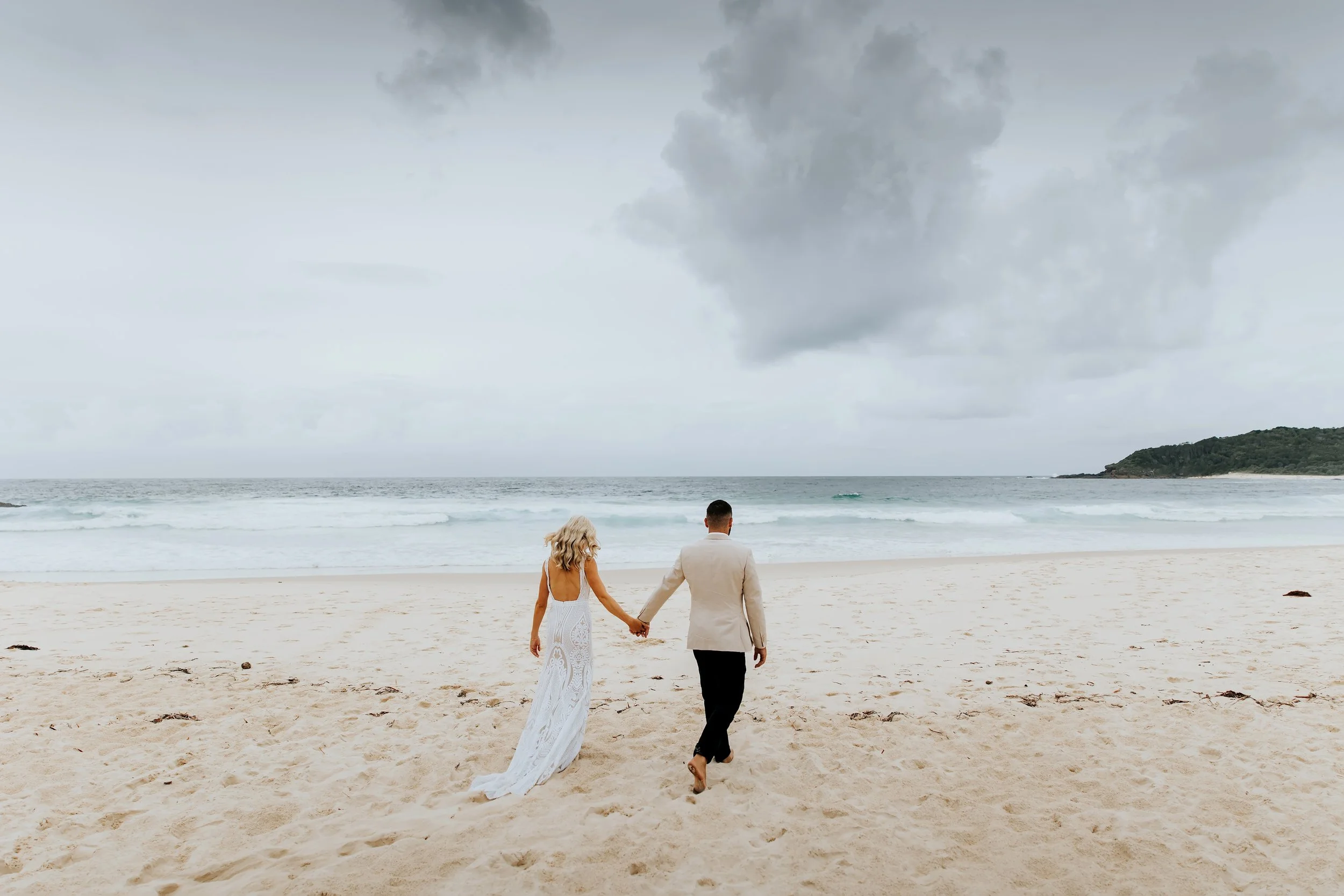 A bride and groom walking hand in hand on the beach under a cloudy sky, with ocean waves and a distant landmass in the background.