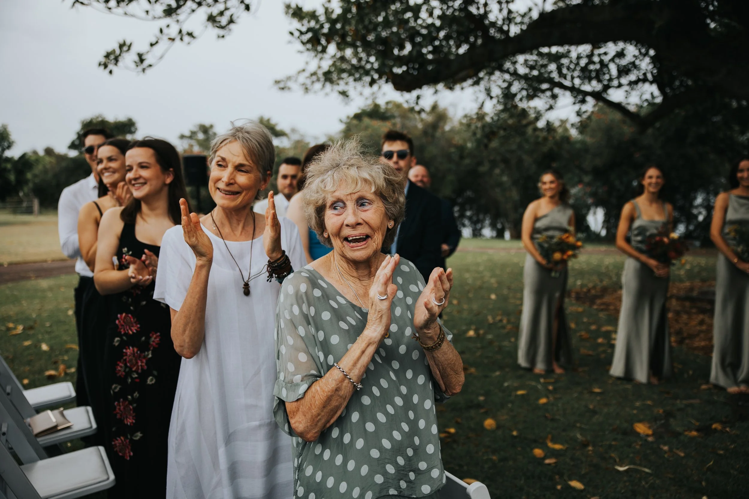 Group of women smiling and clapping outdoors during daytime event, with some women dressed in summer attire, and trees in background.