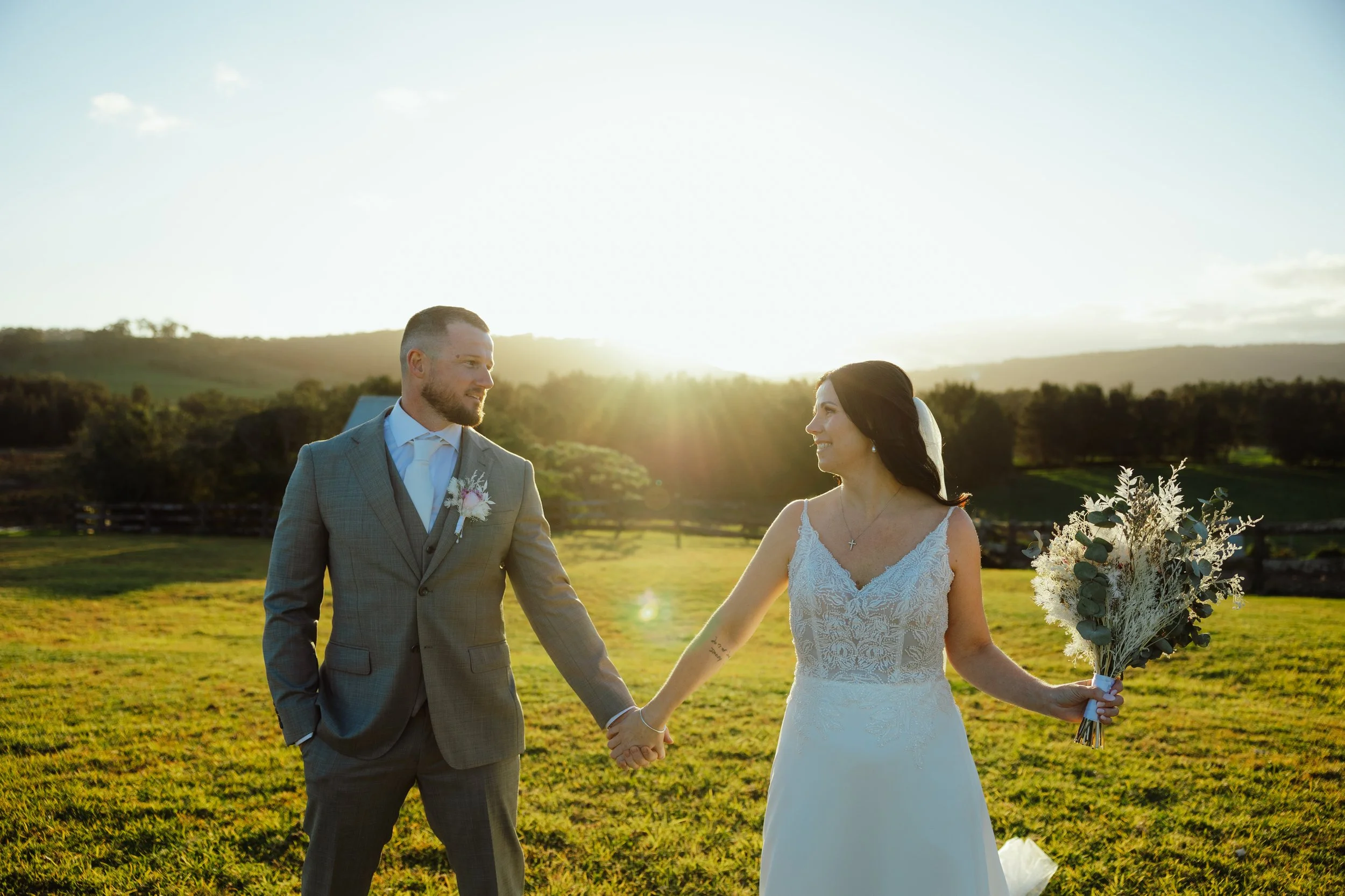 A bride and groom holding hands outdoors during sunset, with a scenic landscape in the background, both smiling at each other.
