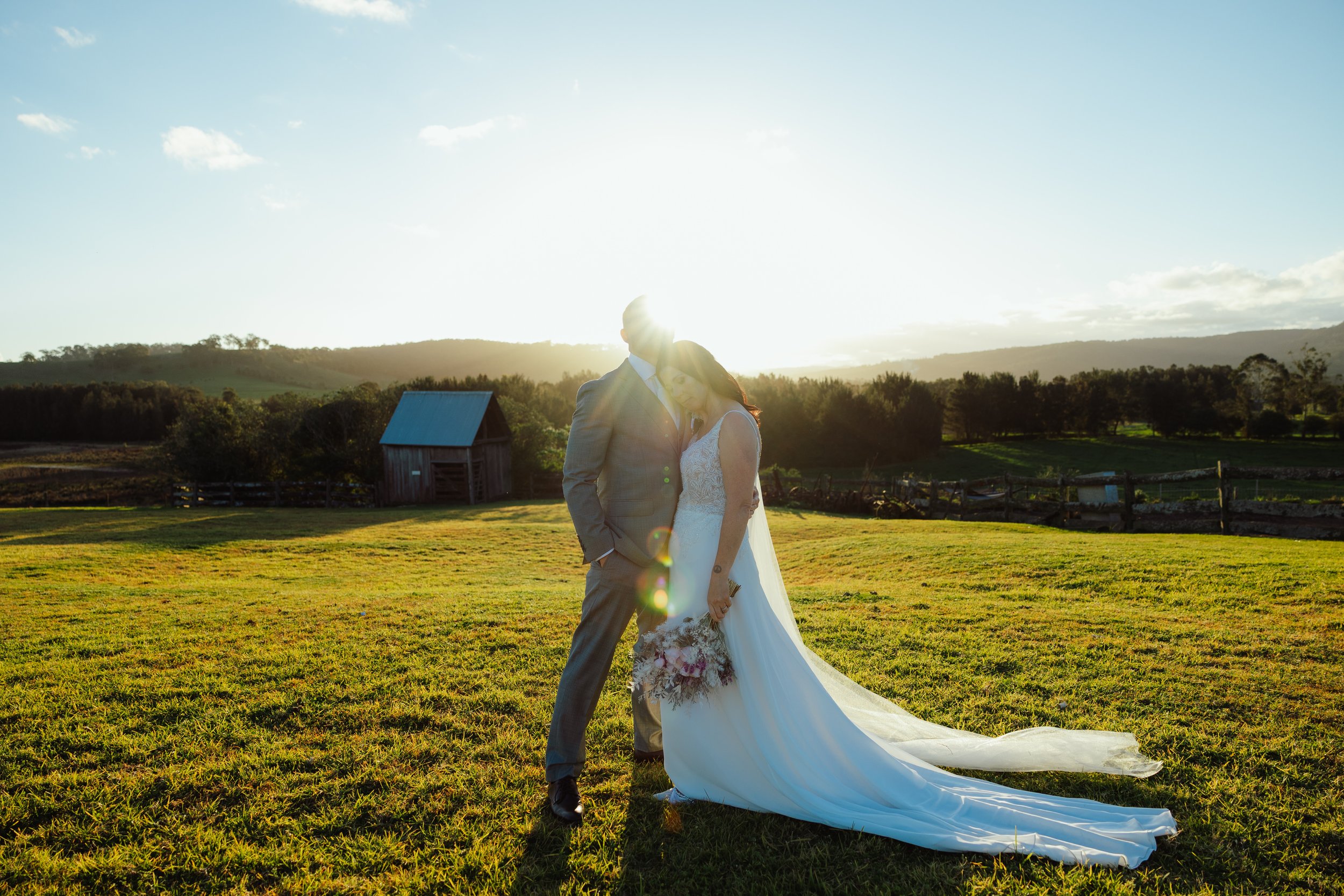 A bride and groom standing together outdoors during sunset, with the bride leaning her head on the groom's shoulder. The bride is wearing a white wedding dress with lace details and holding a bouquet. The groom is wearing a gray suit with a white shi