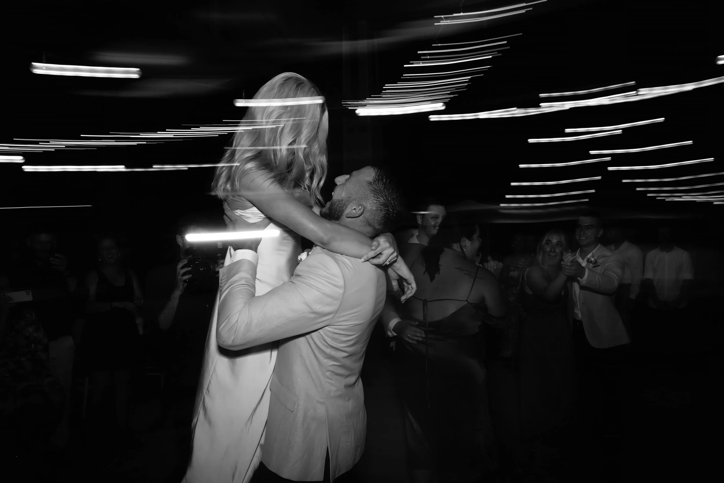 A couple dancing at a celebration with string lights overhead, surrounded by smiling guests.