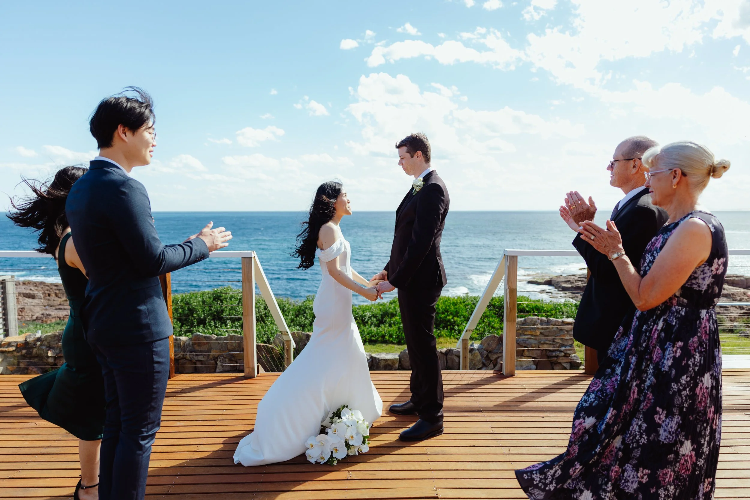 A wedding ceremony taking place outdoors by the ocean. The bride and groom are holding hands and facing each other, surrounded by four people, two on each side, possibly witnesses or officiants. The scene is sunny with a clear sky and gentle waves in
