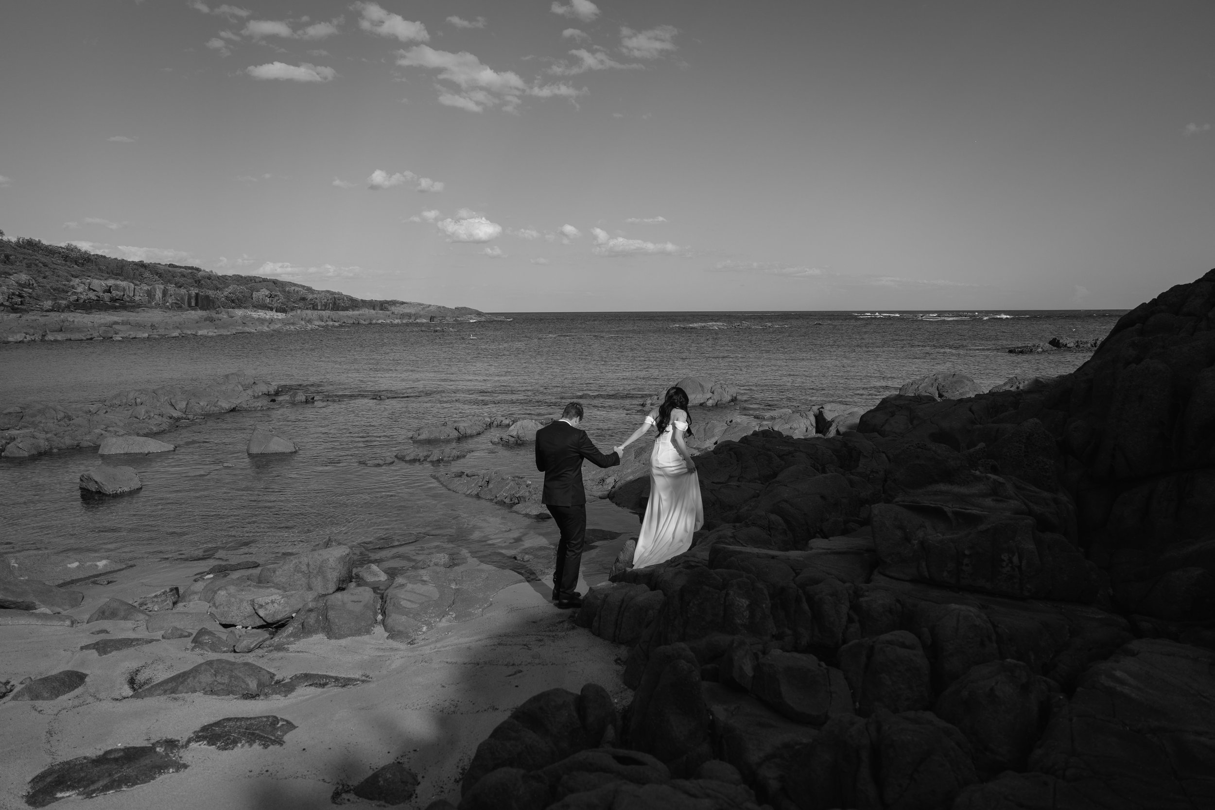 A black and white photograph of a couple holding hands on a rocky beach, with the ocean and a distant shoreline in the background, under a partly cloudy sky.