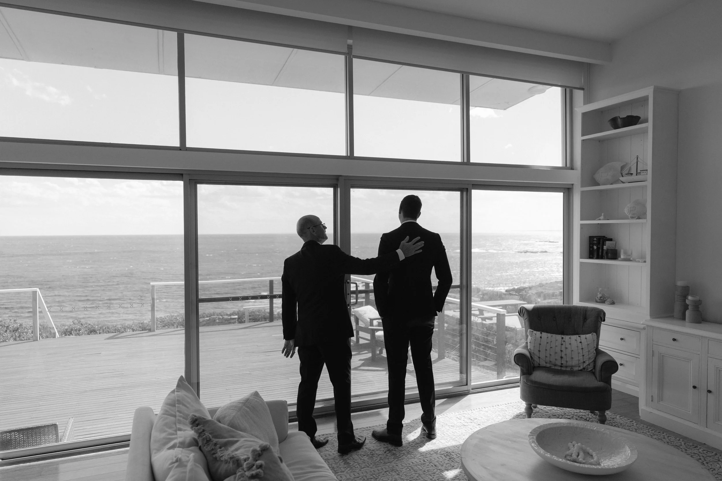Two men in suits stand inside a modern living room, with one placing his hand on the other's shoulder, facing a large window overlooking the ocean. The room has a white bookshelf and a plush armchair, with a sofa partially visible in the foreground.