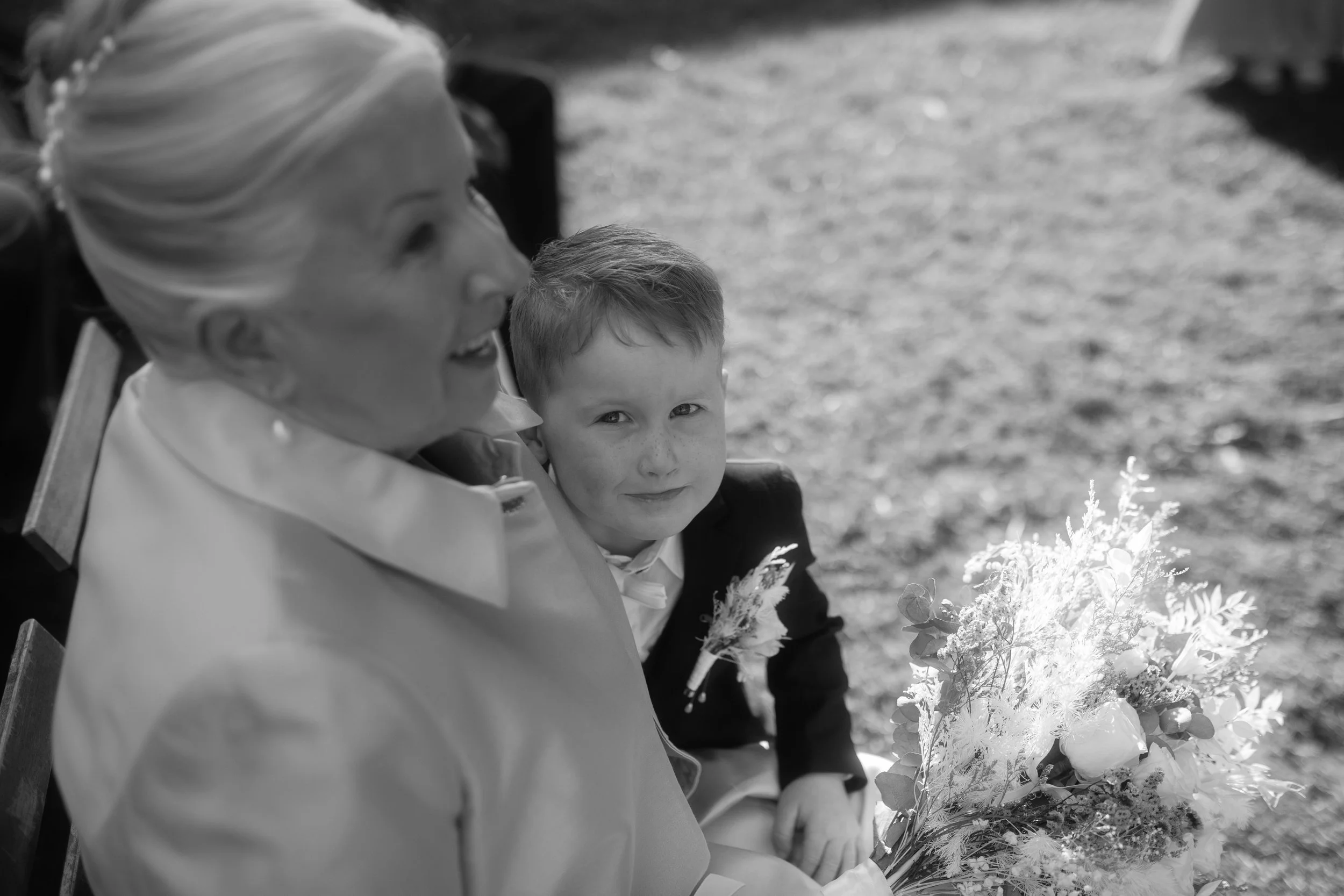 An older woman with light hair, wearing a light-colored blazer, sitting next to a young boy with light hair, wearing a dark jacket, both holding flowers, outdoors on a sunny day.