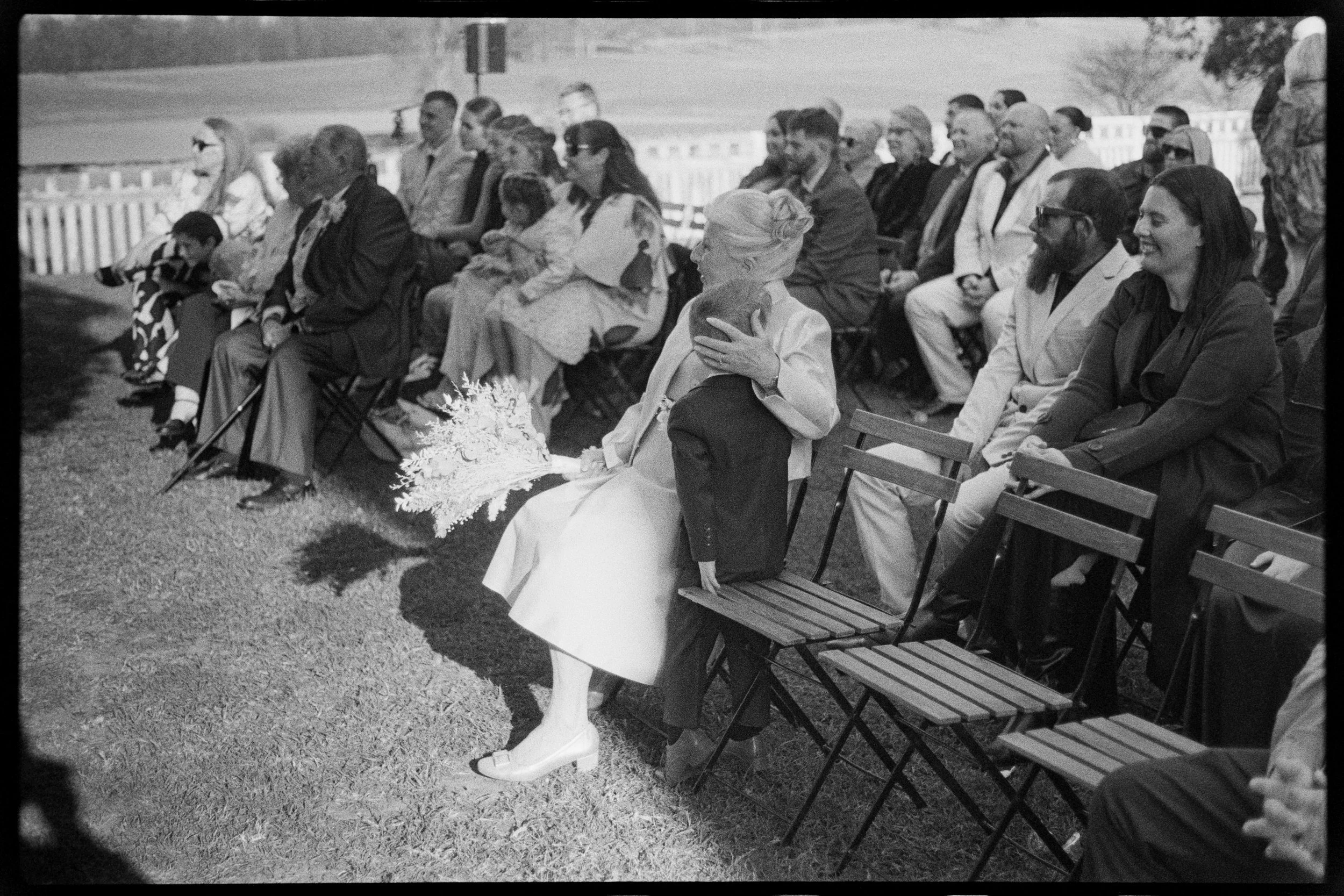 Black and white photograph of a group of people seated outdoors on folding chairs, attending an event. A woman in a light-colored dress is holding a bouquet of flowers, and a young girl is sitting on a man's lap. Others are dressed in formal attire, 