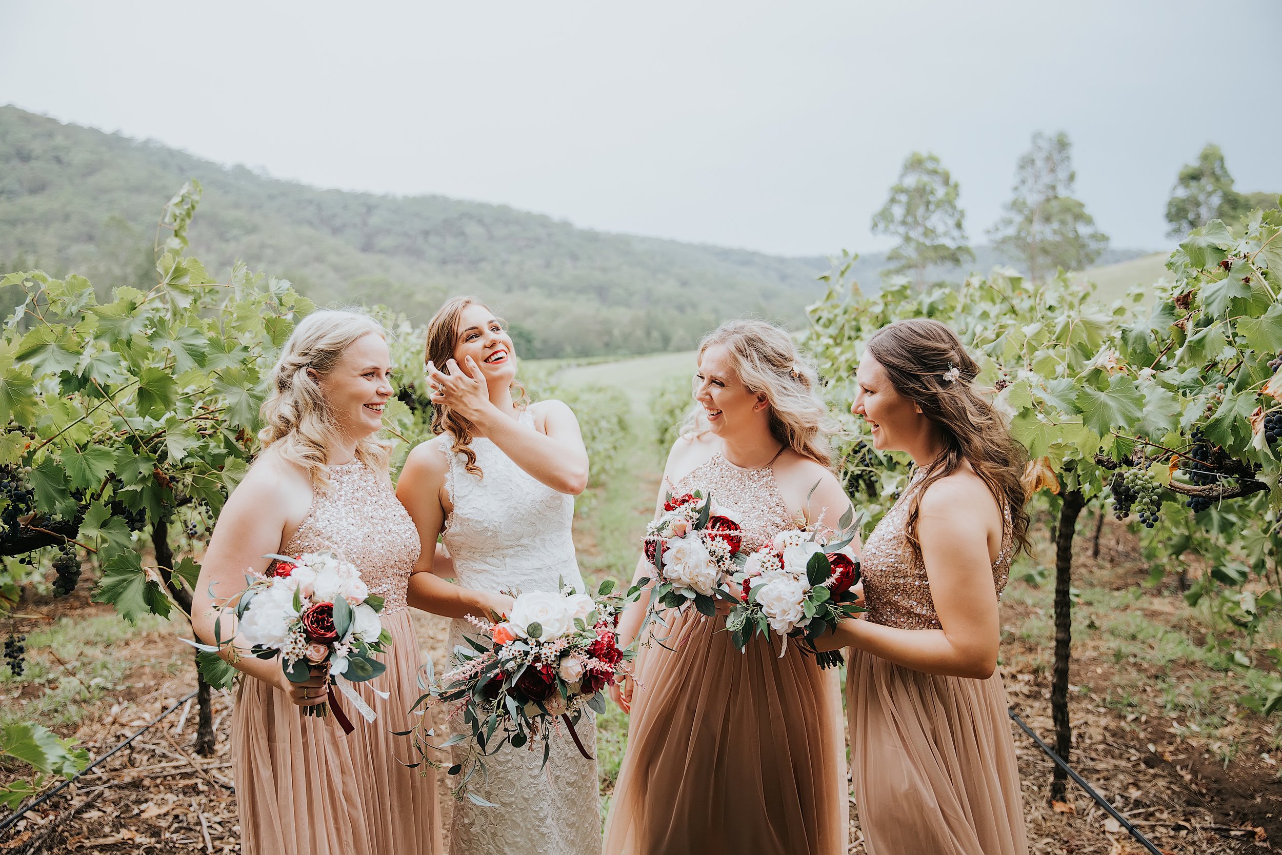 A bride in a white lace dress with four bridesmaids in matching pink sequin tops and pleated skirts, standing in a vineyard with green grapevines and hills in the background, smiling and holding bouquets.