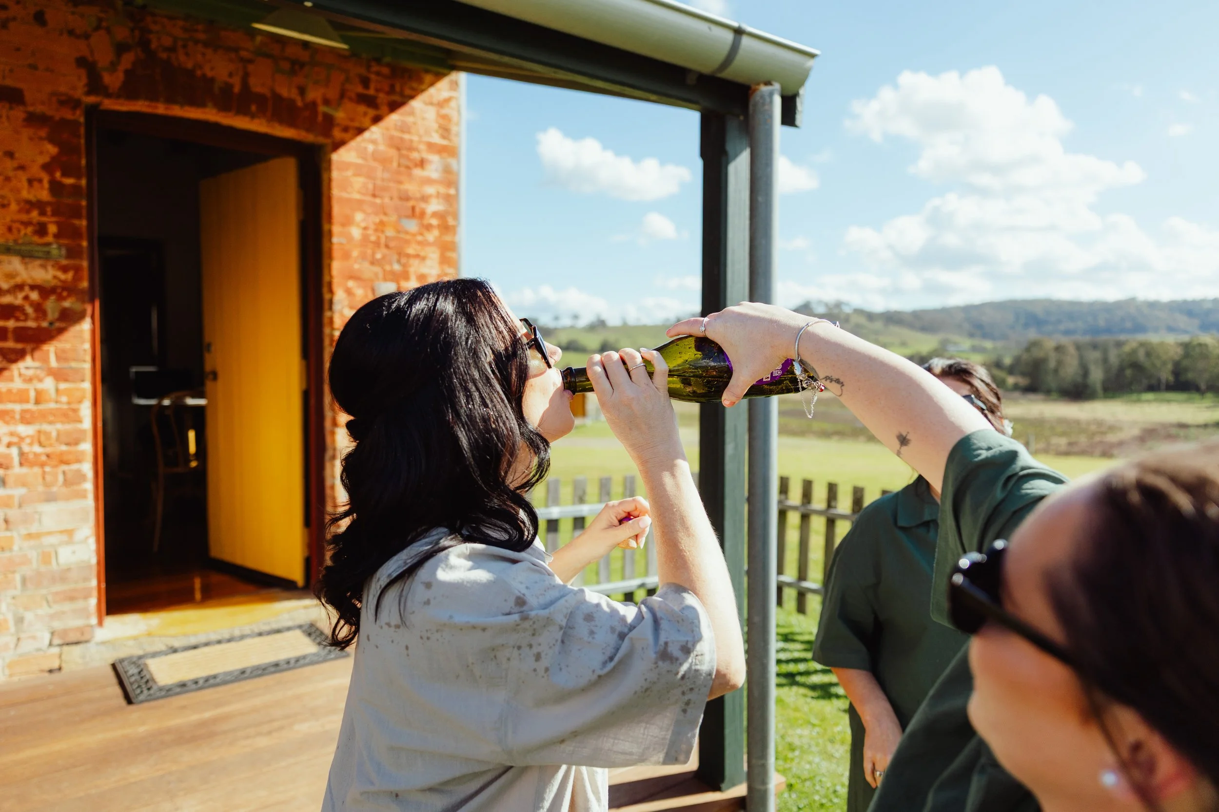 A woman with dark hair and sunglasses drinks a bottle of beer outdoors, with a group of people nearby, at a rural location with a brick building, open door, and green landscape.