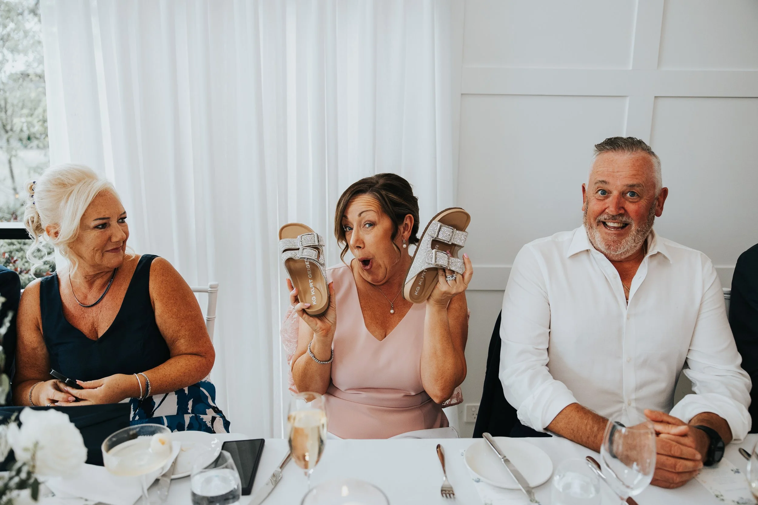 Three adults sitting at a table during a celebration, with one woman excitedly holding up a pair of glittery sandals. The table has food, drinks, and utensils, and the scene is bright with white curtains in the background.
