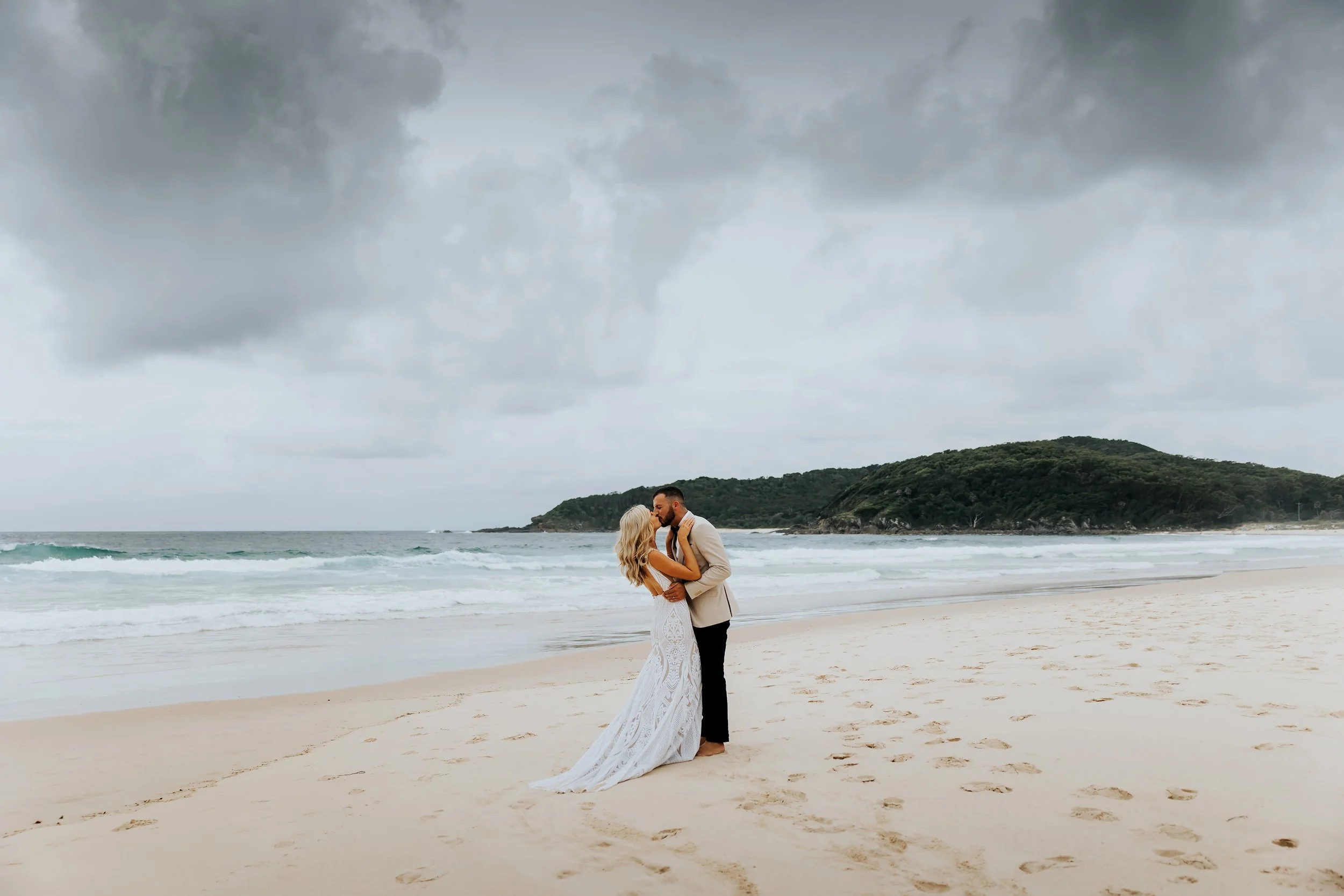 A couple, the bride in a white lace wedding dress and the groom in a beige suit, share a kiss on the beach with gray stormy clouds overhead and an island in the background.