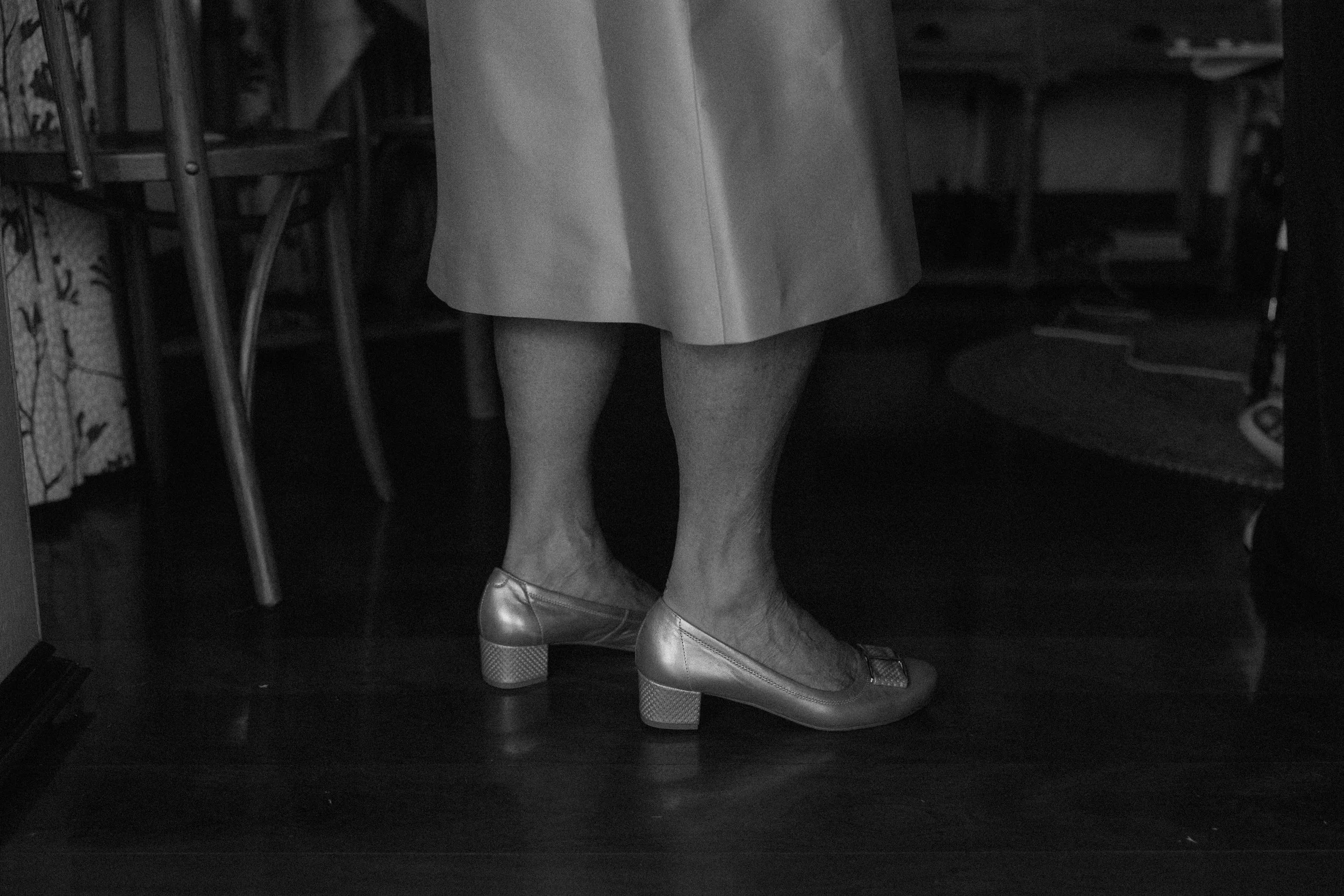 The photo shows the legs and feet of a person wearing a skirt, tights, and high heels, standing on a wooden floor near a chair and a table in a dimly lit room.