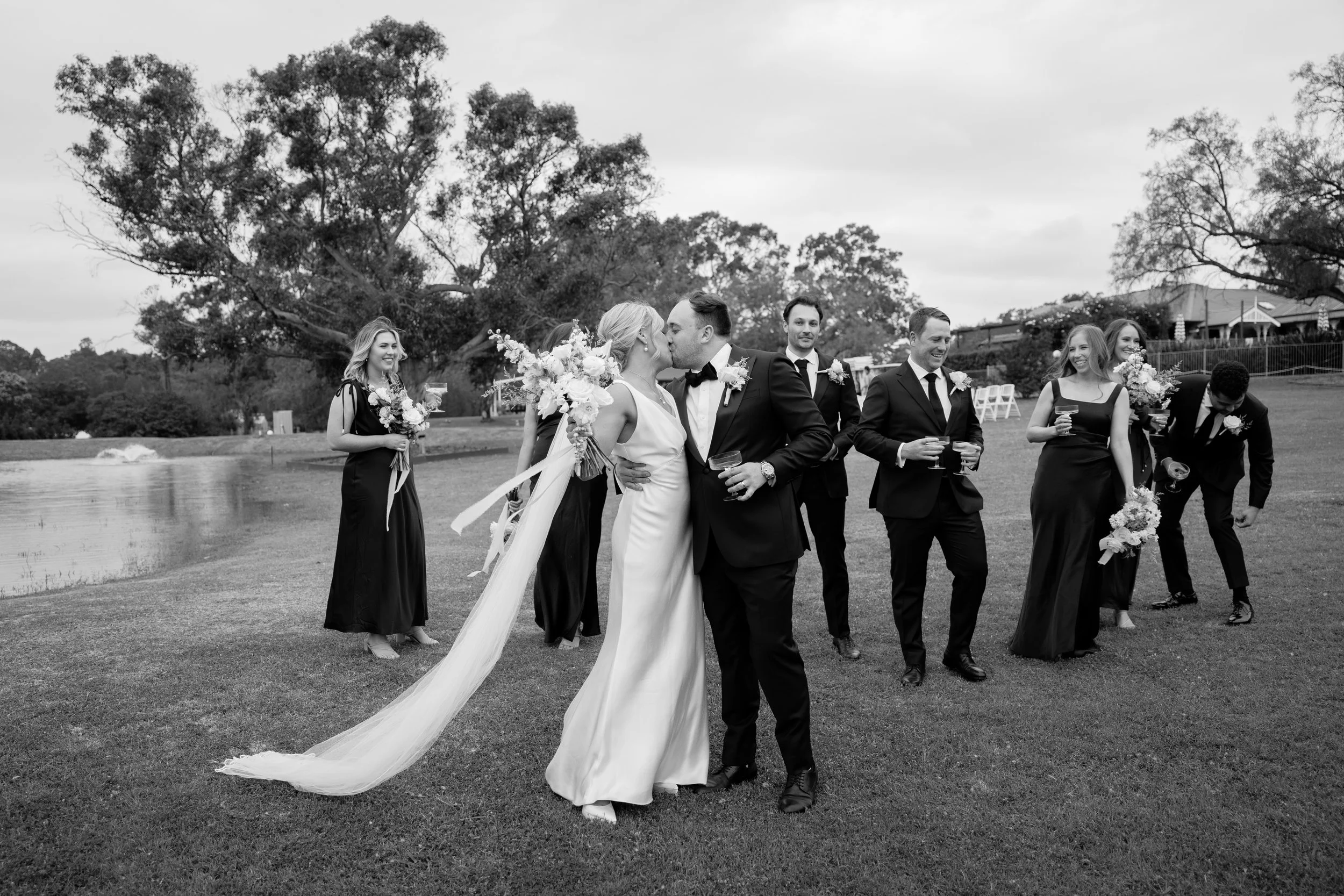 A black and white photo of a wedding scene outdoors with a bride and groom kissing, surrounded by bridesmaids and groomsmen holding flowers and glasses, with trees and a pond in the background.