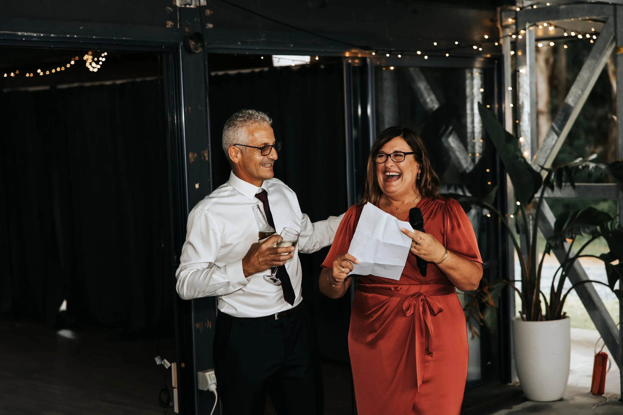 A man and a woman are at a celebration, smiling and holding drinks. The woman is holding a microphone and reading from a piece of paper. They are dressed formally, with the woman in a satin dress and the man in a white shirt with a tie. The setting i