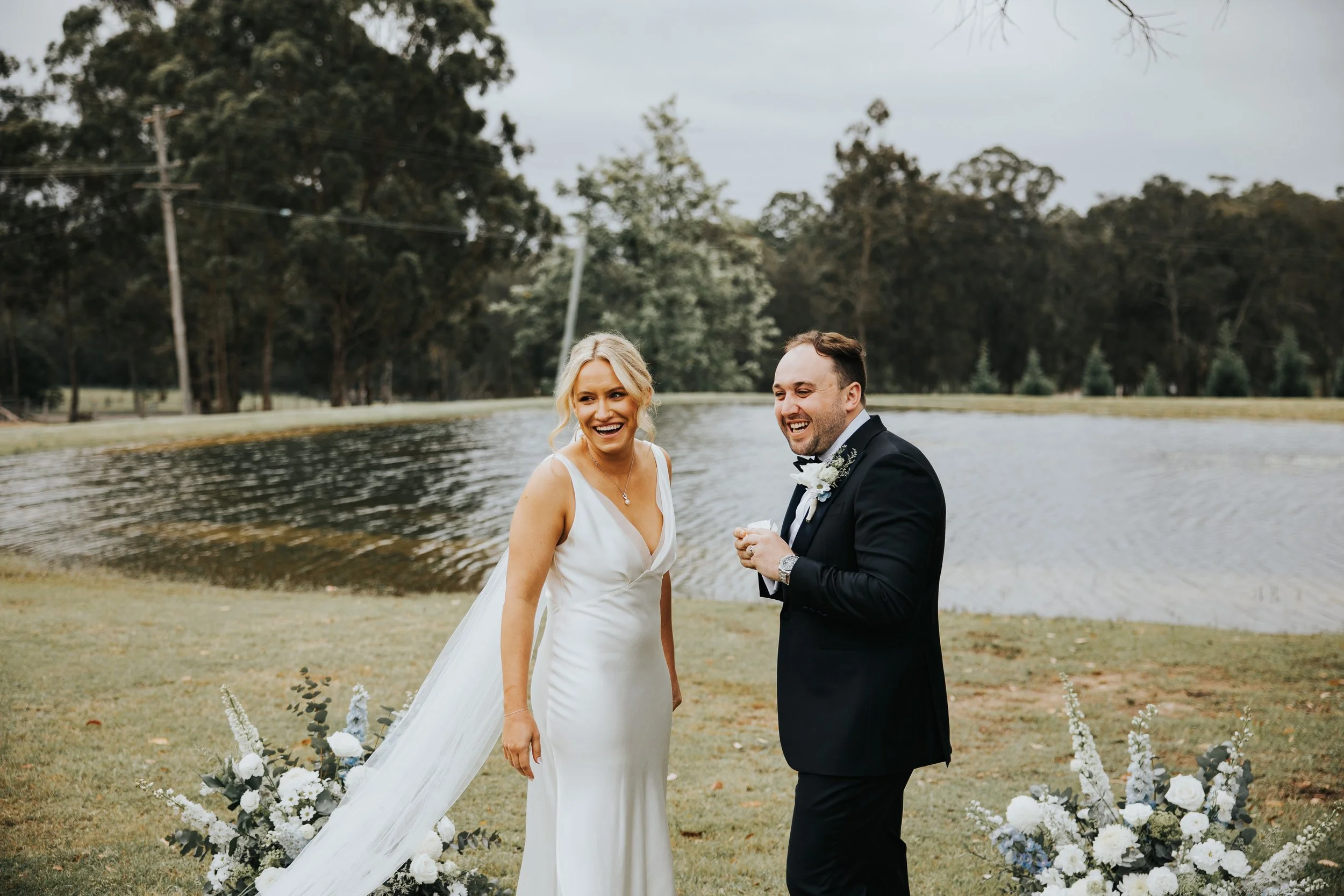 A bride and groom smiling and holding hands during their outdoor wedding ceremony near a lake, with trees and flowers in the background.