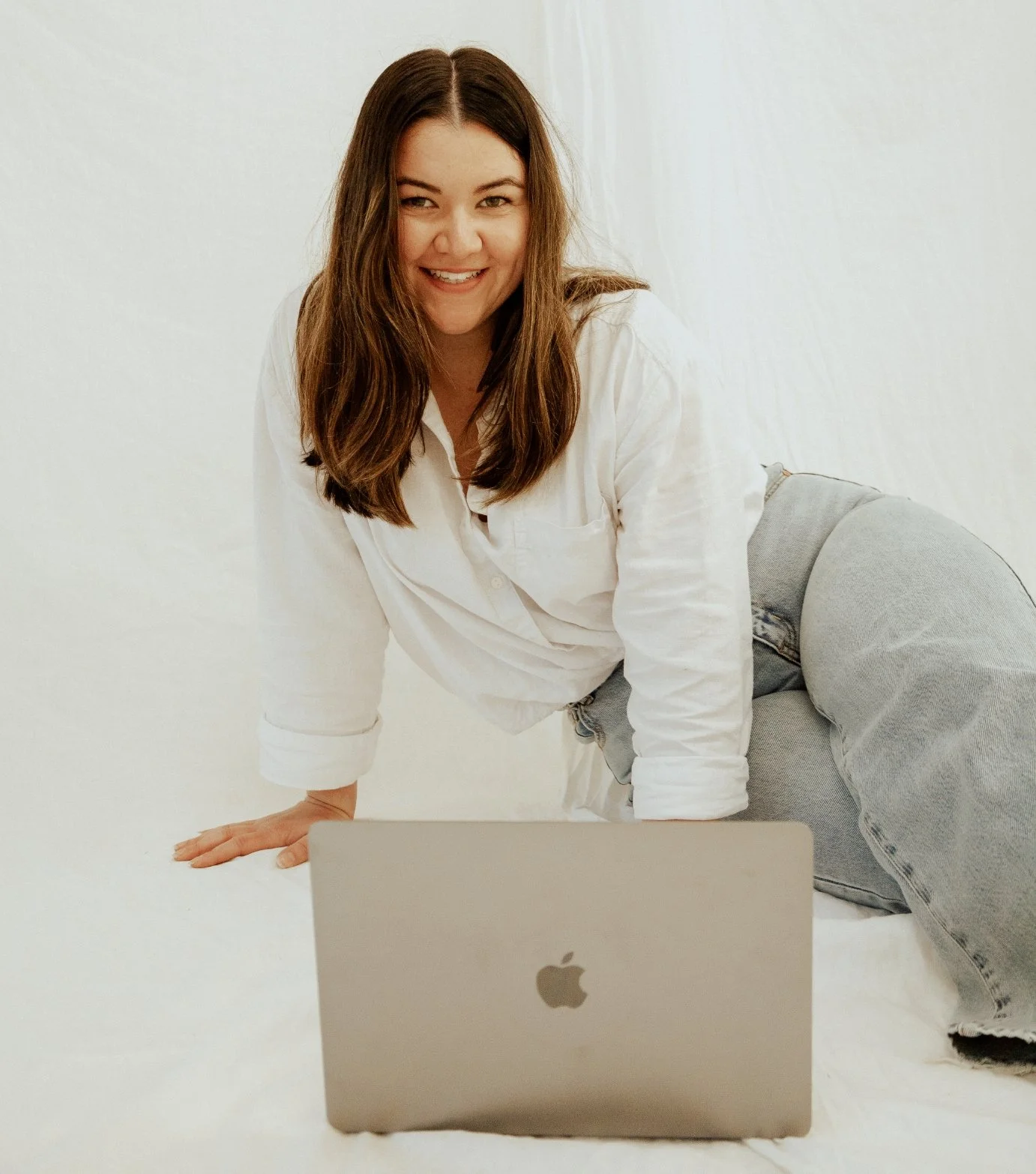 A woman with brown hair and a white shirt, smiling while sitting on a white surface with a silver laptop in front of her.