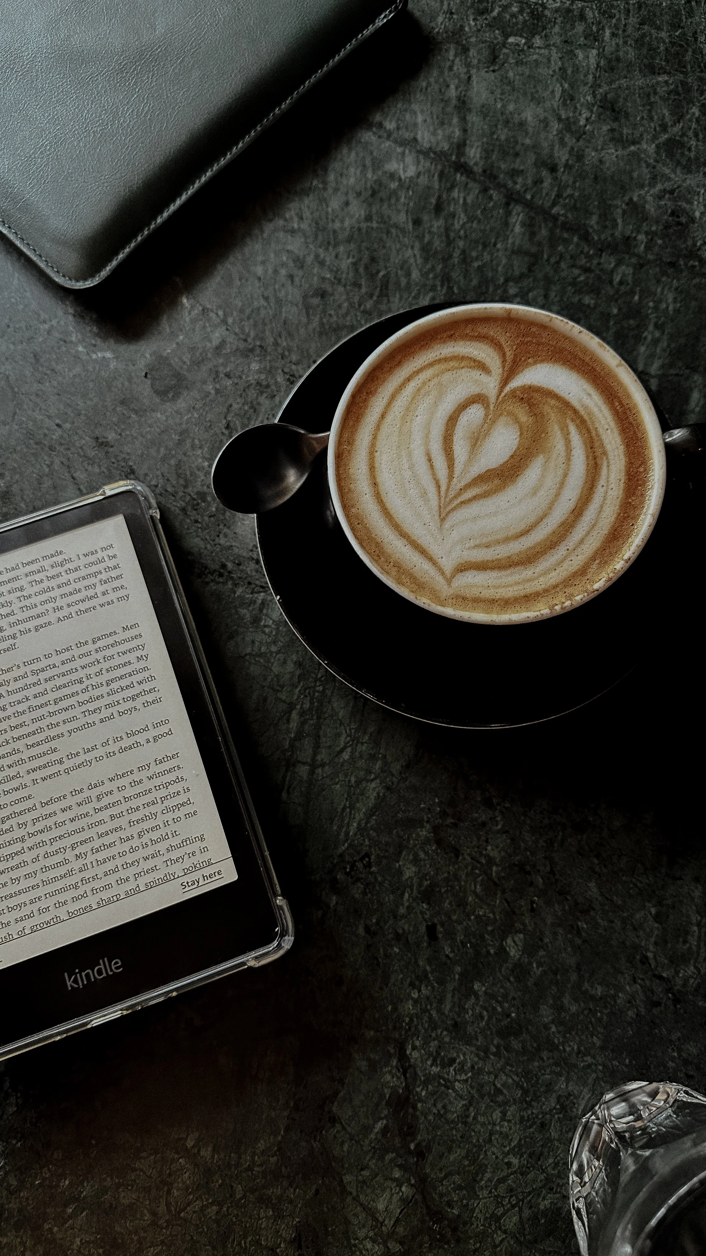 A cup of latte with heart-shaped latte art on a black saucer, a small spoon on the saucer, an e-reader displaying text, and a glass of water on a dark textured table.
