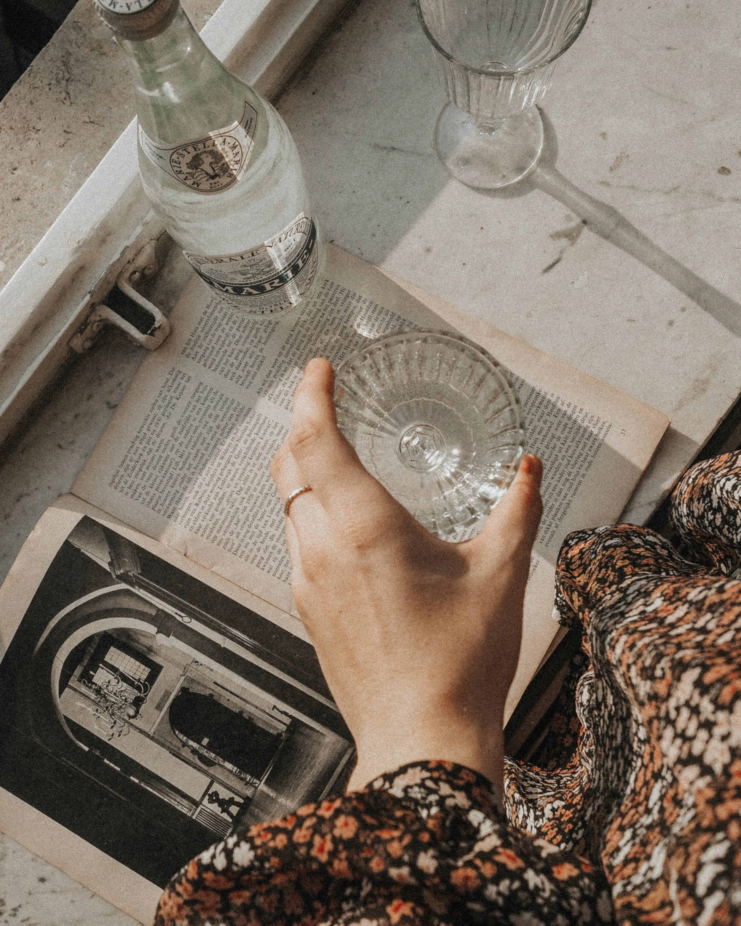A person holding a glass of water above an open book on a table. There is a bottle of San Pellegrino sparkling water and a wine glass on the table. The person wears a floral-patterned sleeve and a ring on their finger.