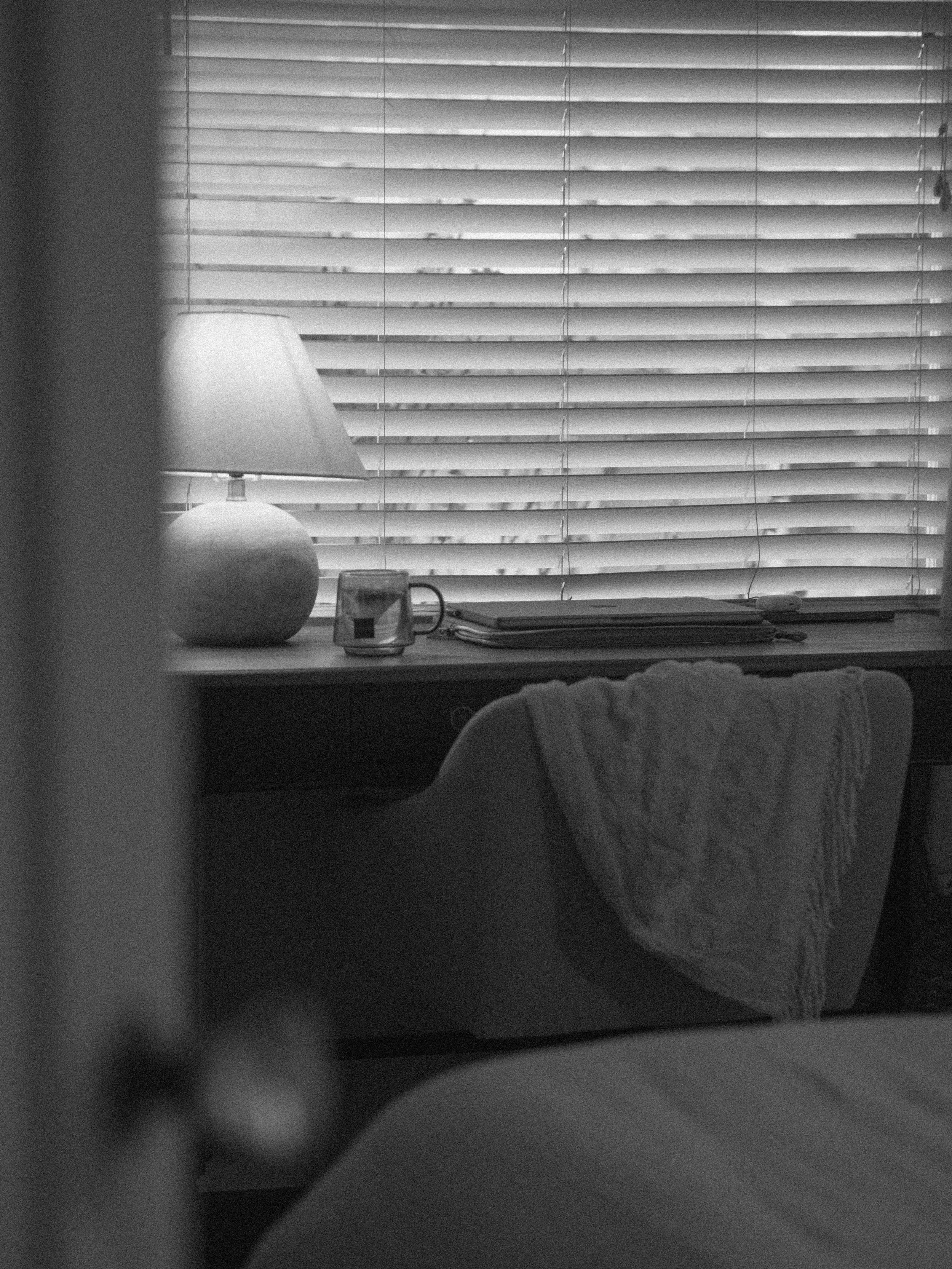 A black and white photo of a windowsill with blinds, a table lamp, a mug, some folders, and a blanket draped over the back of a chair.