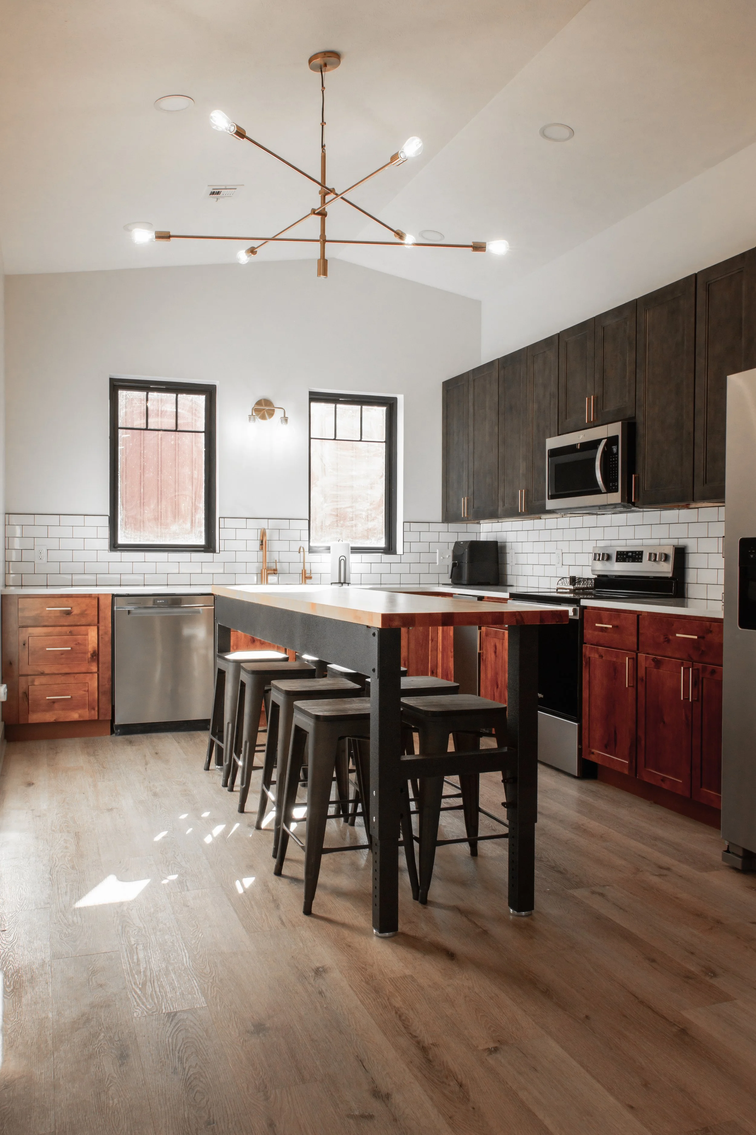 Modern kitchen with two black framed windows, white subway tile backsplash, wooden cabinets, stainless steel appliances, and a black kitchen island with bar stools. A chandelier with exposed bulbs hangs from the ceiling.