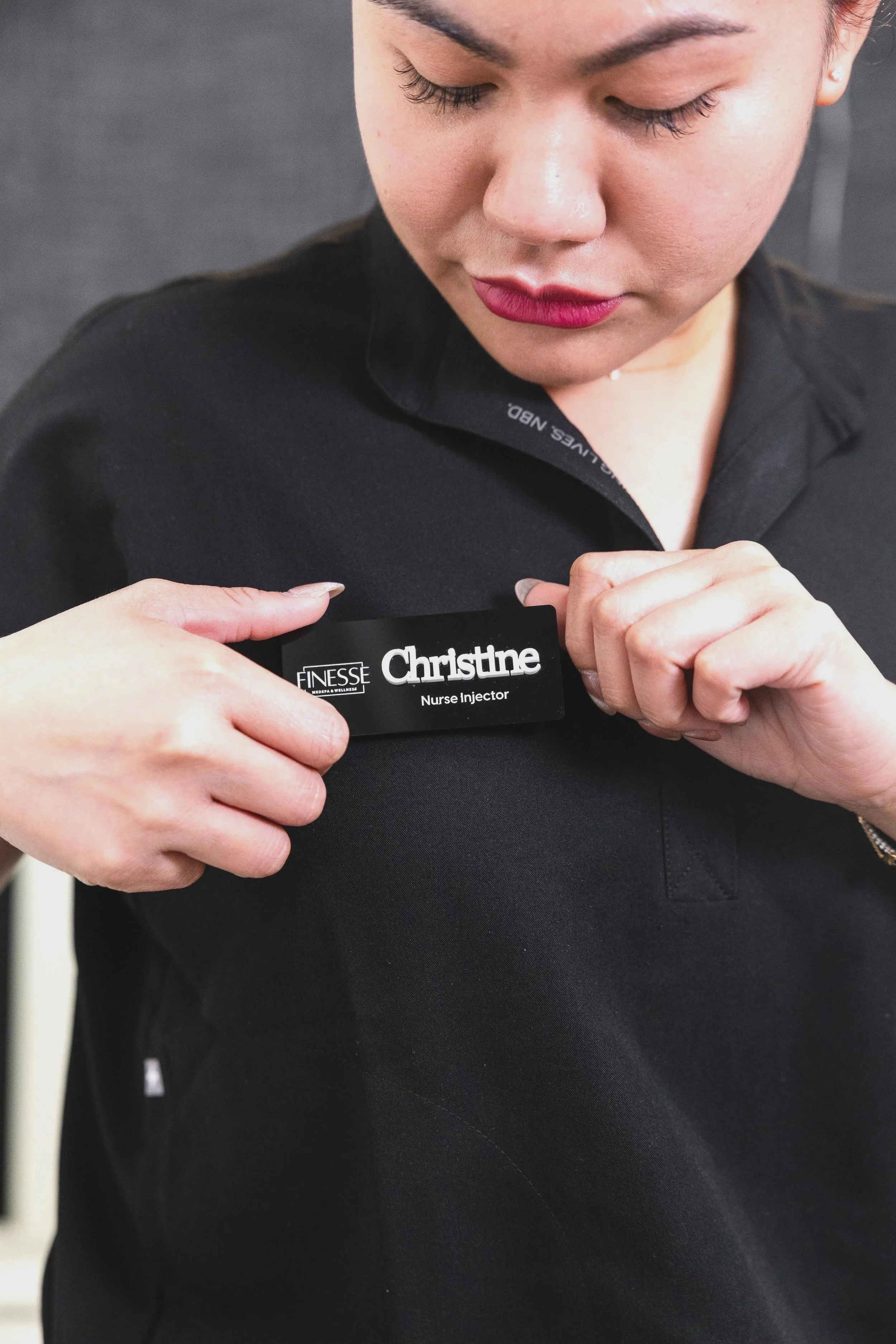 A woman in a black shirt is attaching a name badge that reads 'Christine Nurse Injector' to her shirt.
