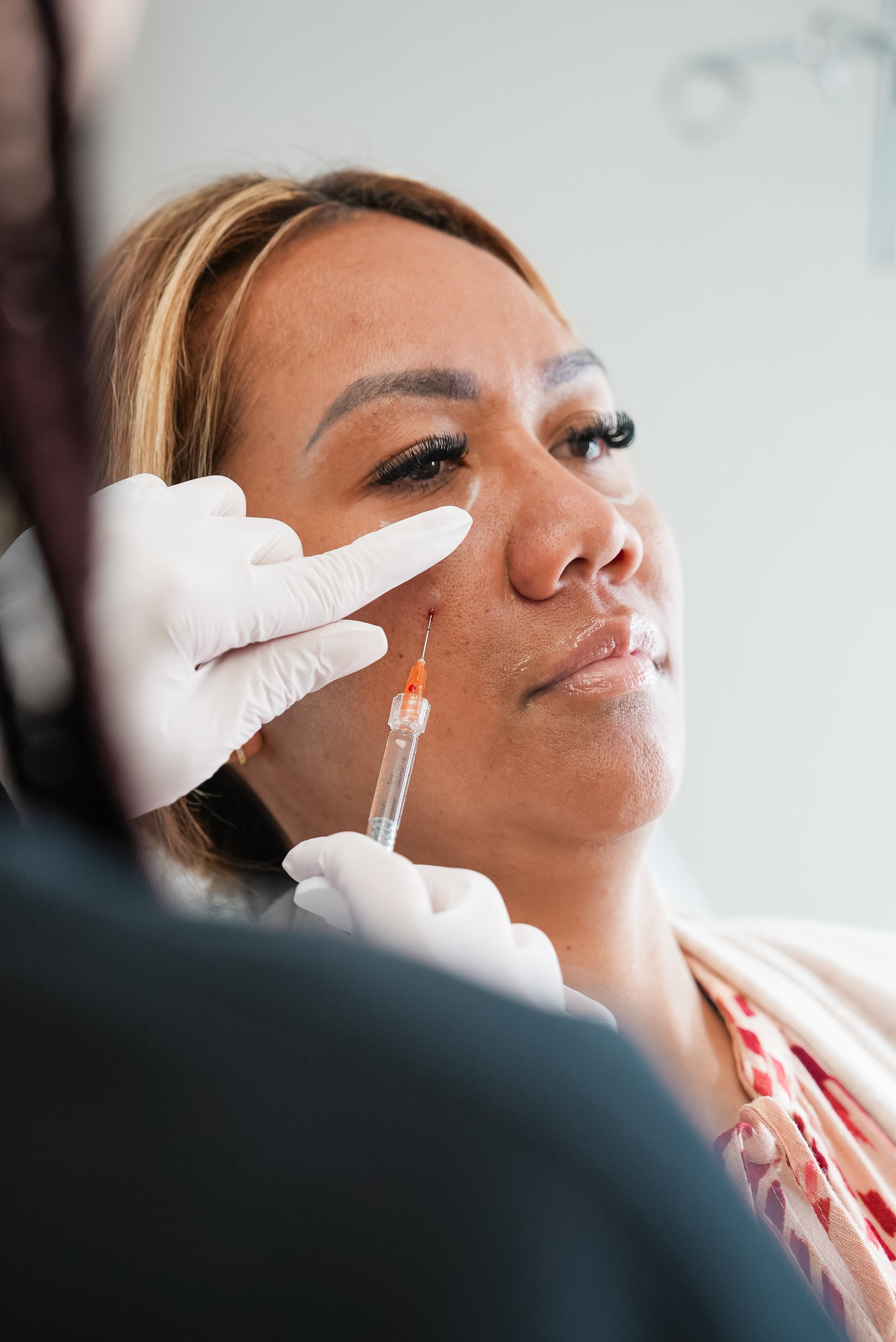A woman receiving a cosmetic treatment with a syringe, while a medical professional wearing white gloves administers an injection near her cheek.