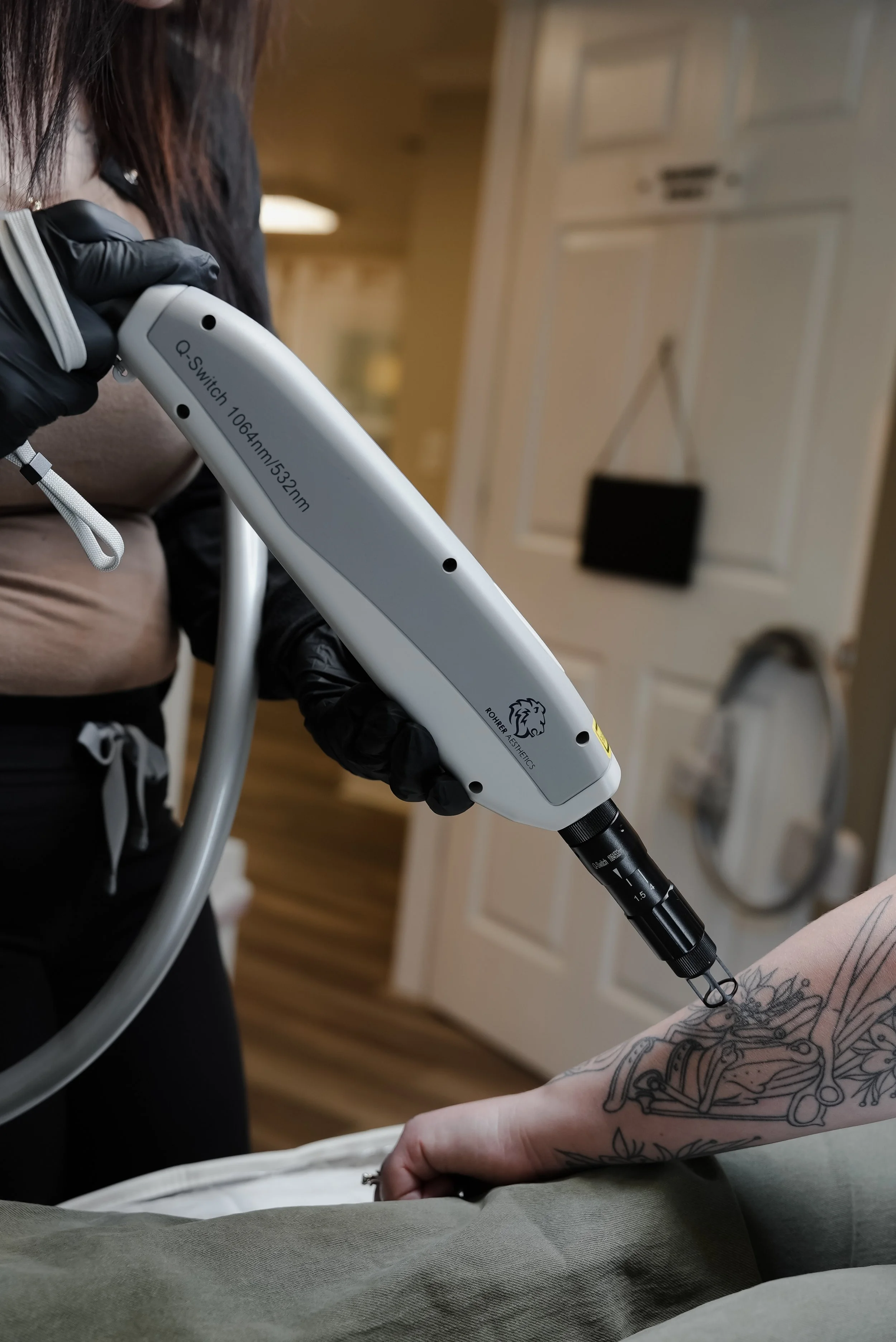 A person wearing black gloves holding a medical or aesthetic device close to a tattooed hand on a bed or chair in a room with wooden floors and a cabinet.