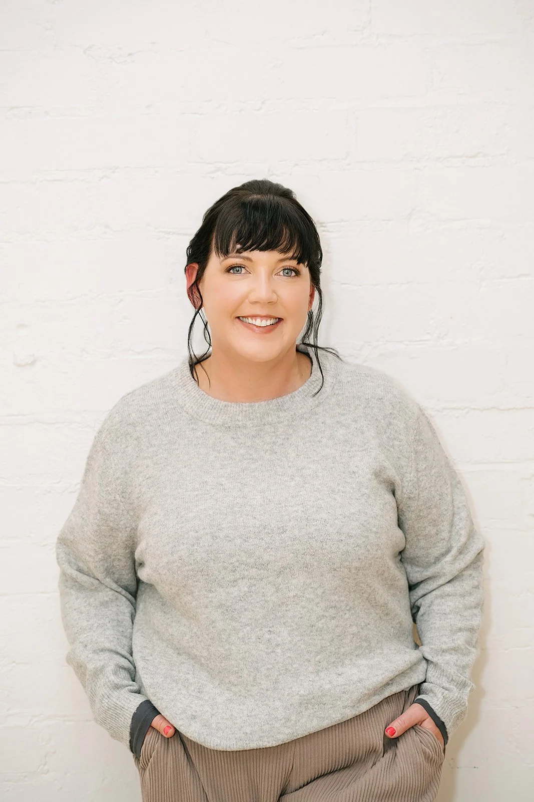 Smiling woman standing against a white brick wall, wearing a grey sweater and relaxed pants with hands in pockets, conveying a confident, approachable, and professional presence