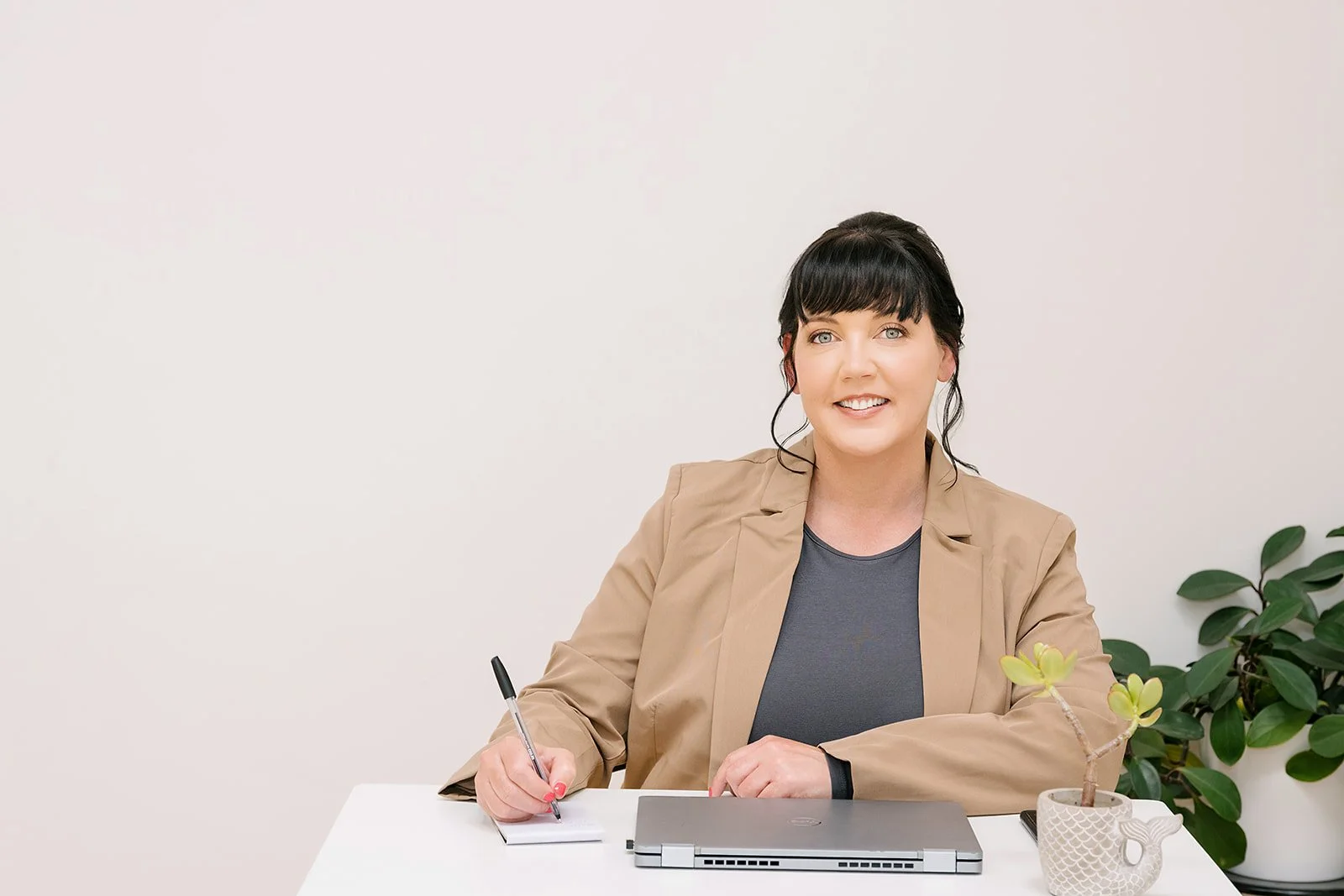 Woman seated at a desk, smiling while writing on a notepad beside a laptop and potted plant, wearing a neutral blazer in a clean, professional workspace, conveying a confident and approachable practice setting.