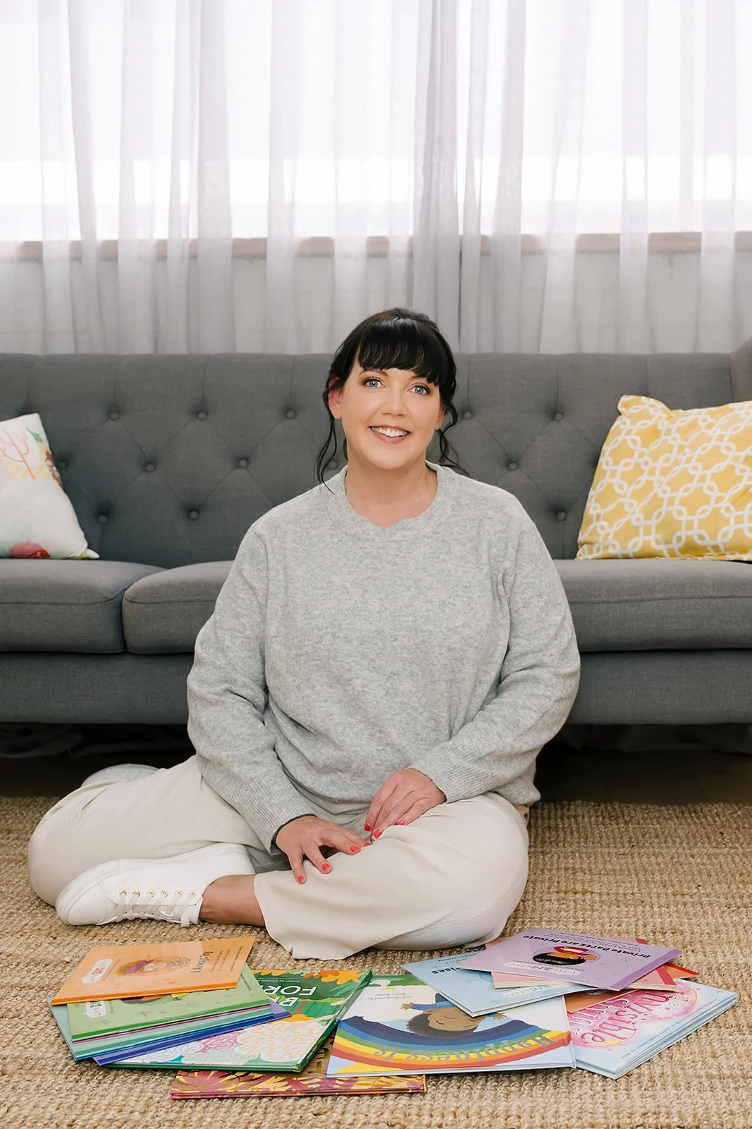 Woman sitting cross-legged on a rug in front of a grey sofa, smiling warmly, wearing a light grey sweater and pale pants, with children’s books spread out in front of her, conveying a calm, supportive, and approachable professional setting.