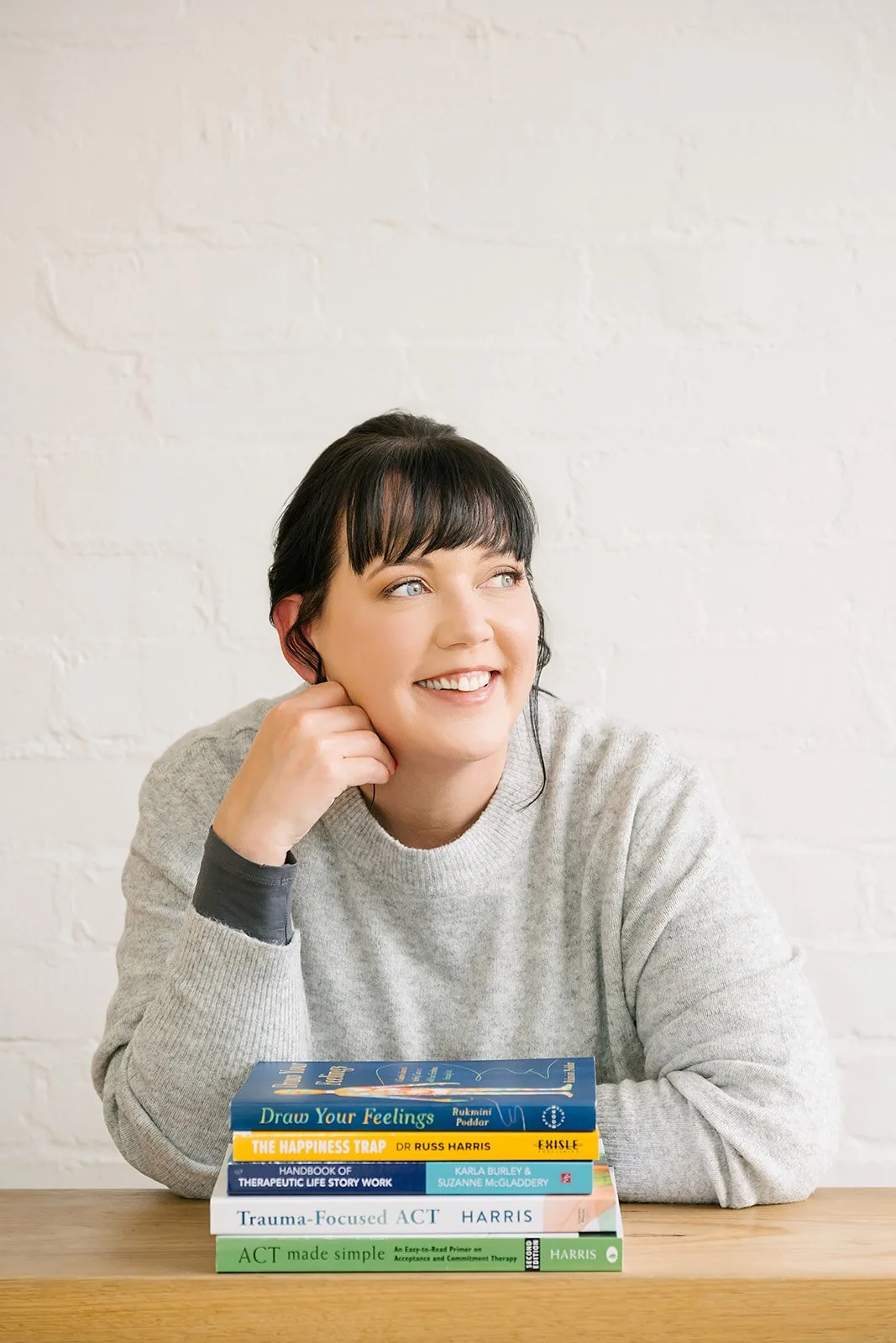 Smiling woman seated at a table, resting her chin on her hand, with a stack of therapy and social work books in front of her, against a white brick wall, conveying a thoughtful, knowledgeable, and approachable professional presence.