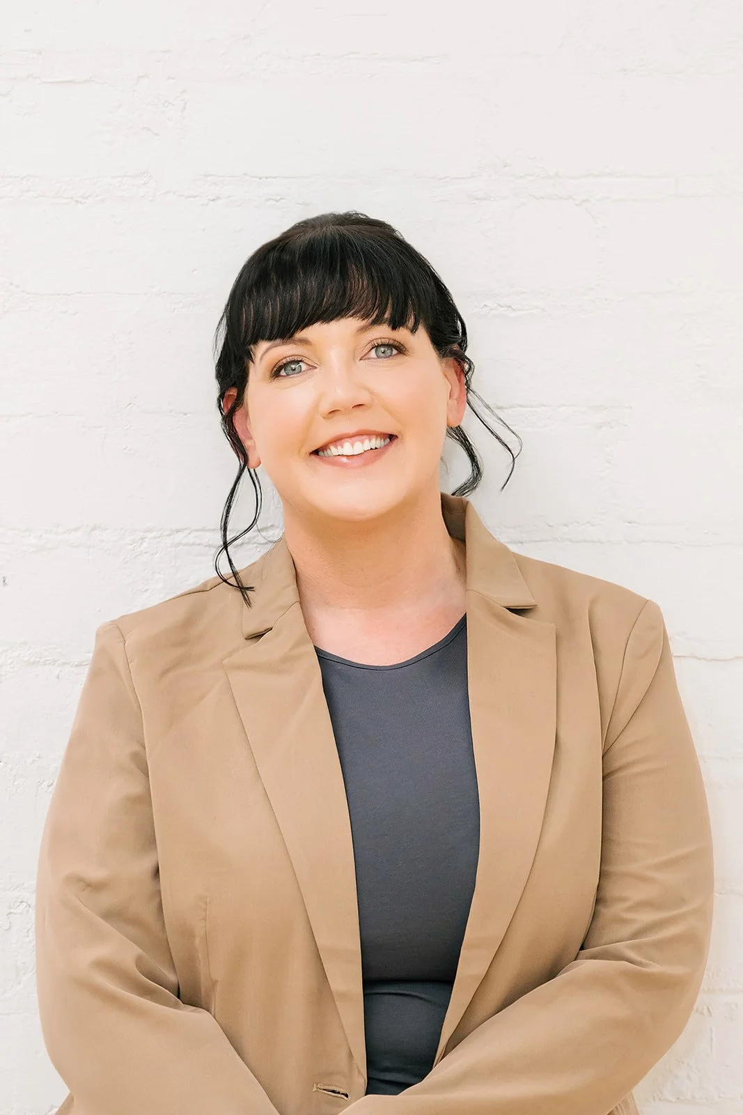 Smiling woman with dark hair and fringe, wearing a tan blazer over a dark top, standing against a white brick wall with a relaxed, approachable posture.