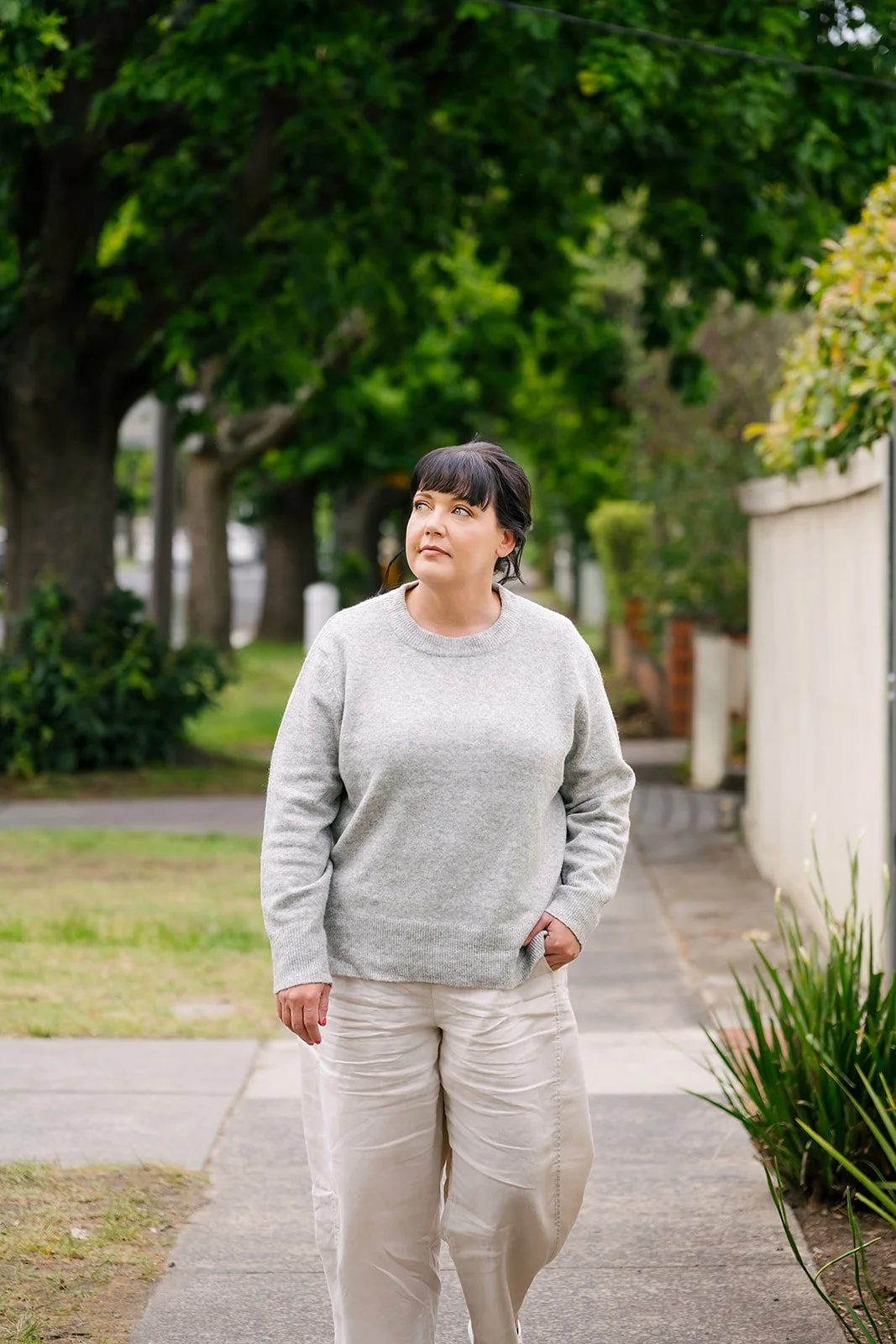 Laura Simpson, social worker and founder of Lets Reseed, walking along a leafy residential footpath, wearing a light sweater and neutral pants, with a calm, reflective expression in a natural outdoor setting.
