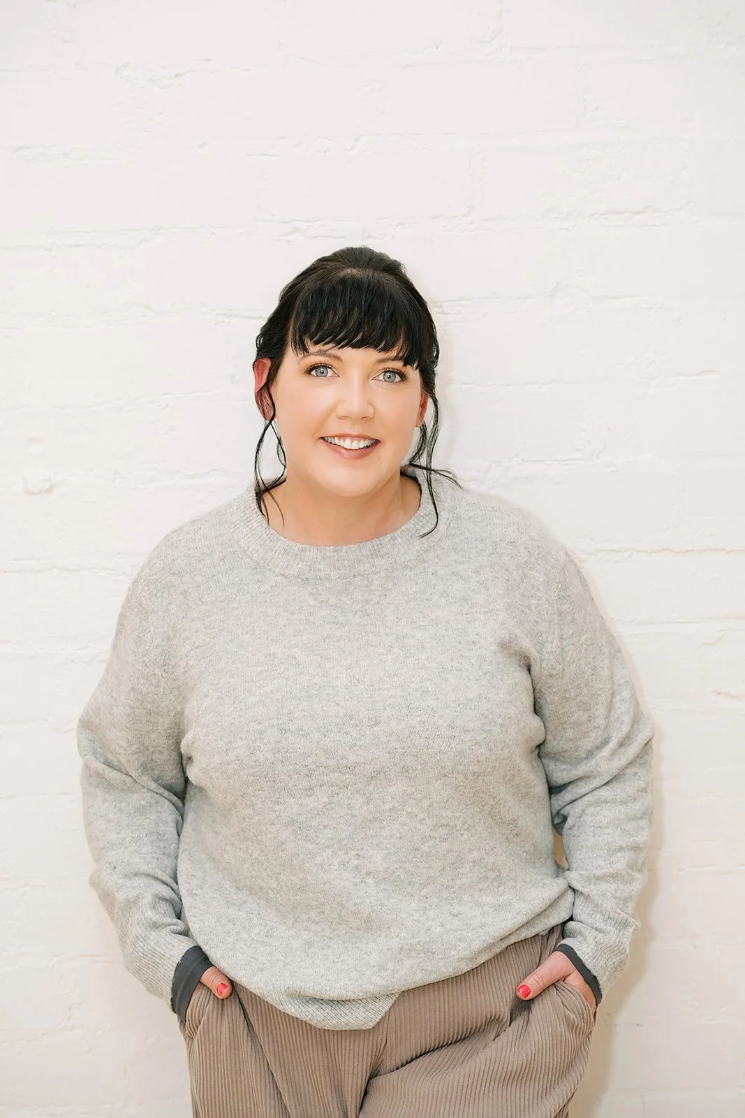 Smiling woman standing against a white brick wall with hands in her pockets, wearing a grey sweater and relaxed pants, conveying a warm, confident, and approachable professional presence