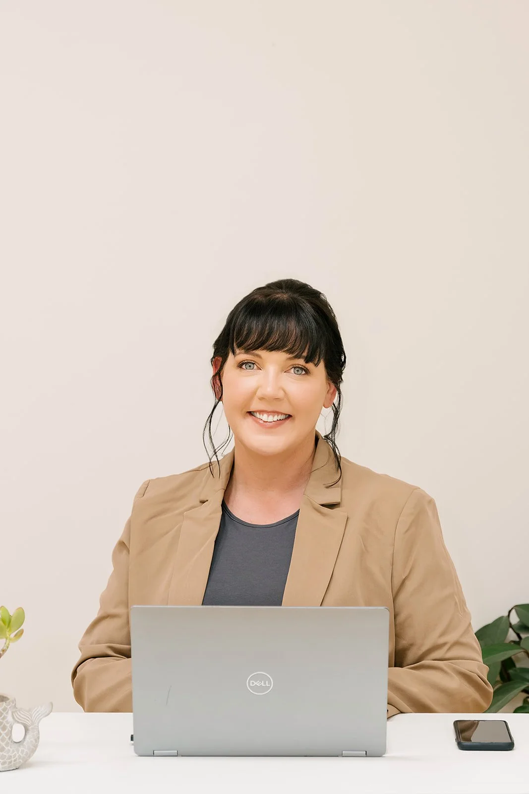 A woman with black hair and bangs, wearing a beige blazer and dark shirt, sitting at a white desk with a laptop and a smartphone, smiling at the camera, against a plain light background.