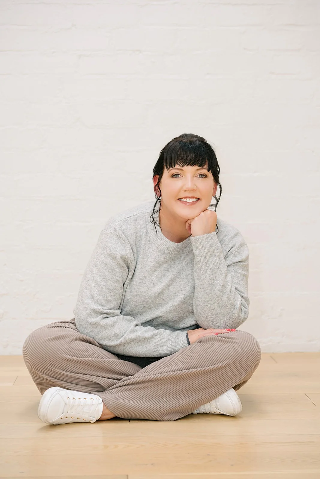 Woman sitting cross-legged on a wooden floor against a white brick wall, smiling warmly, wearing a grey sweater, relaxed pants, and white sneakers, conveying a calm and approachable professional presence.
