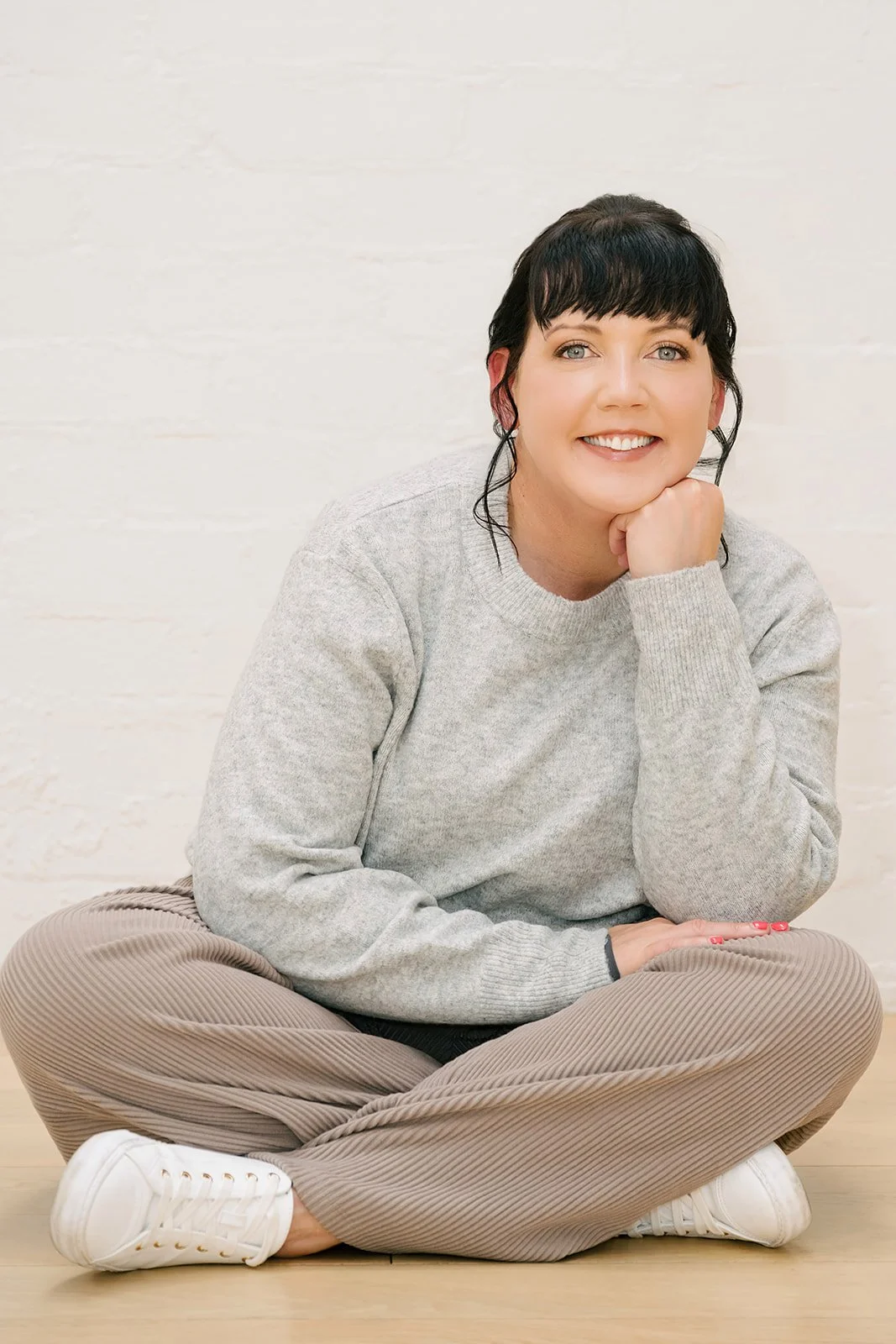 Woman sitting cross-legged on a wooden floor against a white brick wall, smiling gently with her chin resting on her hand, wearing a grey sweater, relaxed pants, and white sneakers, conveying a calm, thoughtful, and approachable professional presence