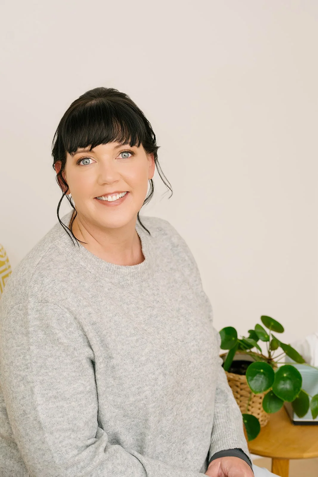 Smiling woman seated in a light, calm office space, wearing a grey sweater, with a green potted plant on a small table beside her, conveying a warm and approachable professional environment.