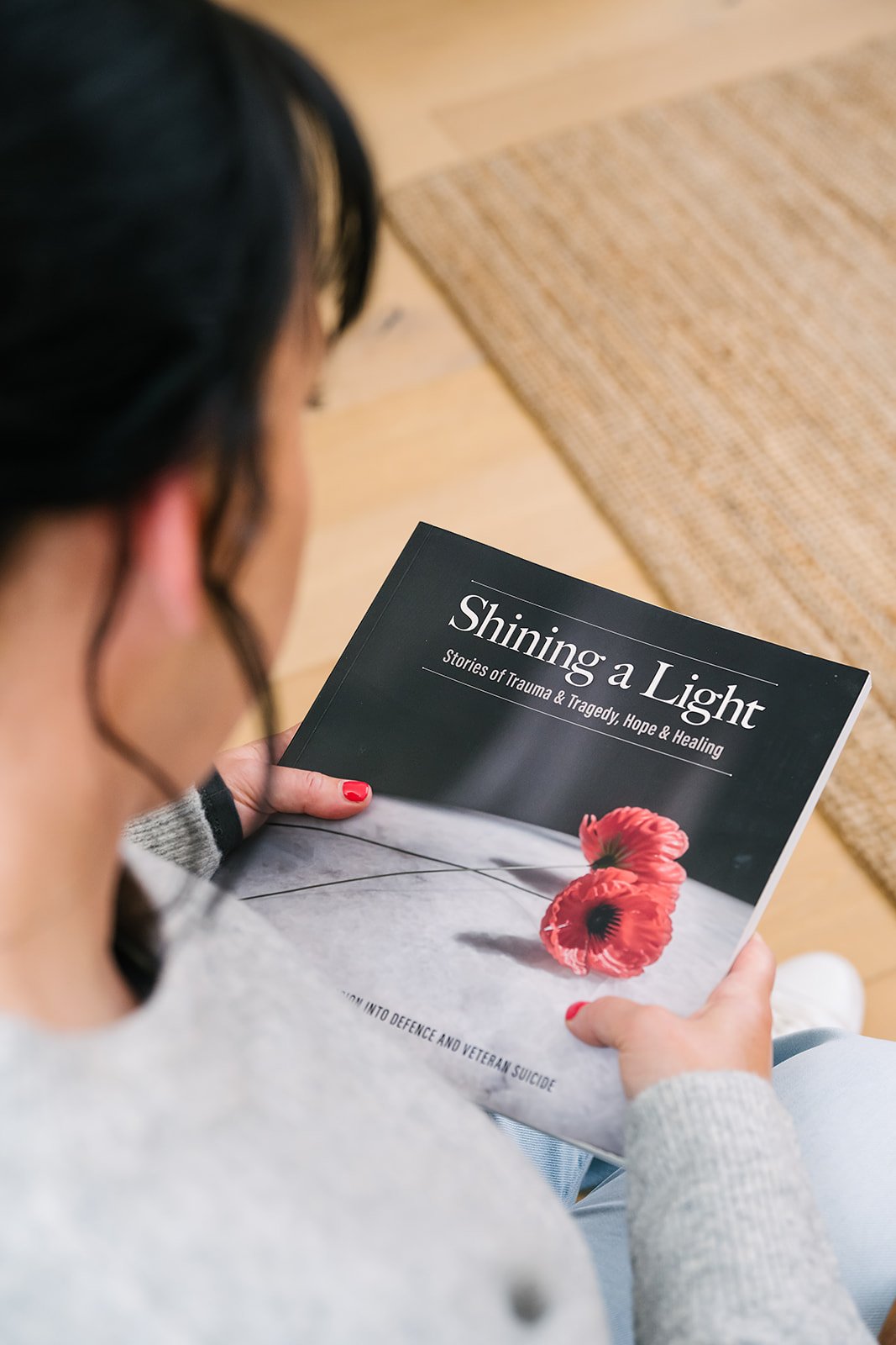 A woman with dark hair and red nails is sitting on the floor and reading a book titled 'Shining a Light: Stories of Trauma & Tragedy, Hope & Healing.' The book's cover features two red poppies lying on a gray surface, with a wooden floor and a woven rug in the background.