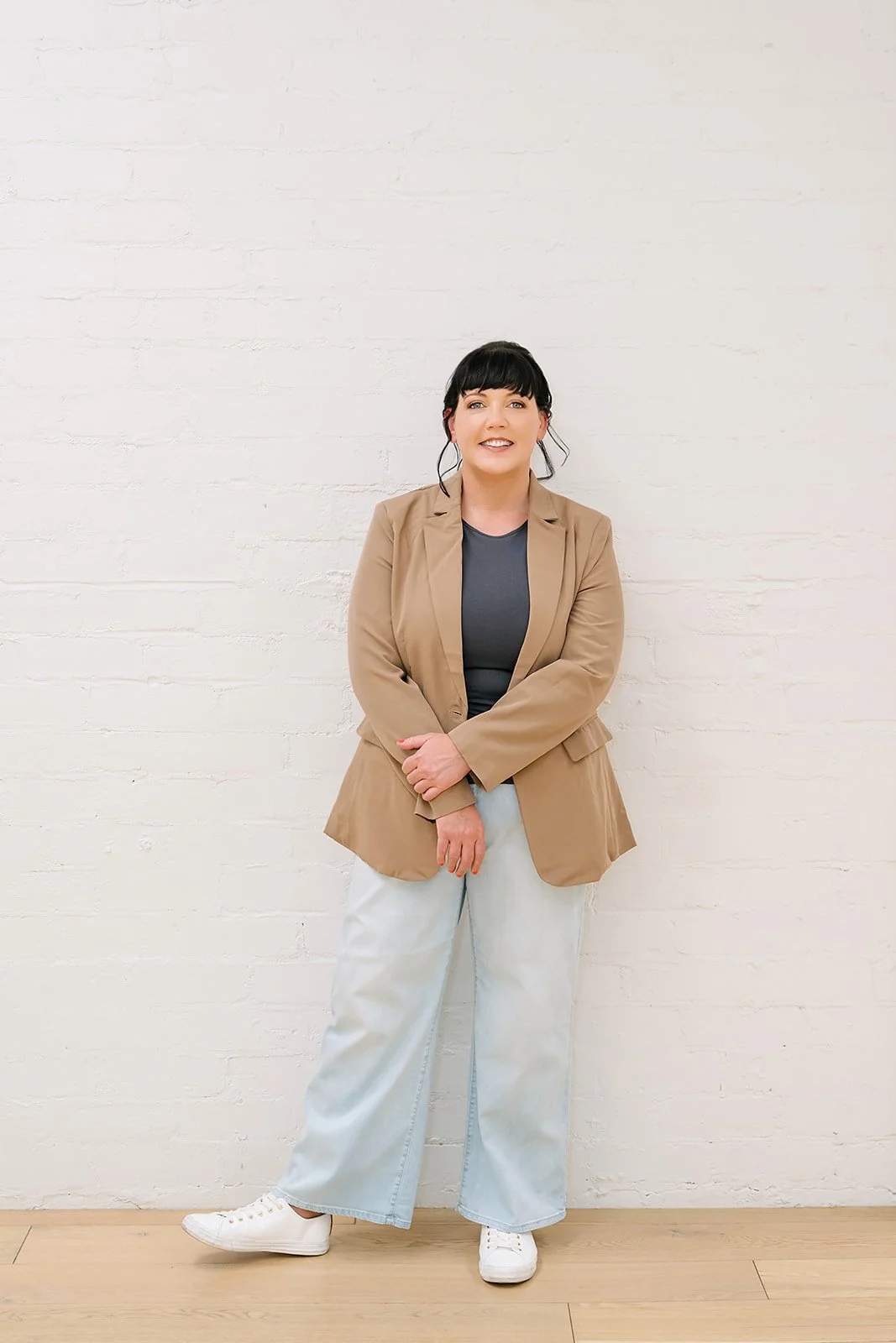 Laura Simpson, social worker and founder of Lets Reseed, standing against a light brick wall, wearing a neutral blazer, casual jeans and sneakers, smiling in a relaxed and approachable pose.