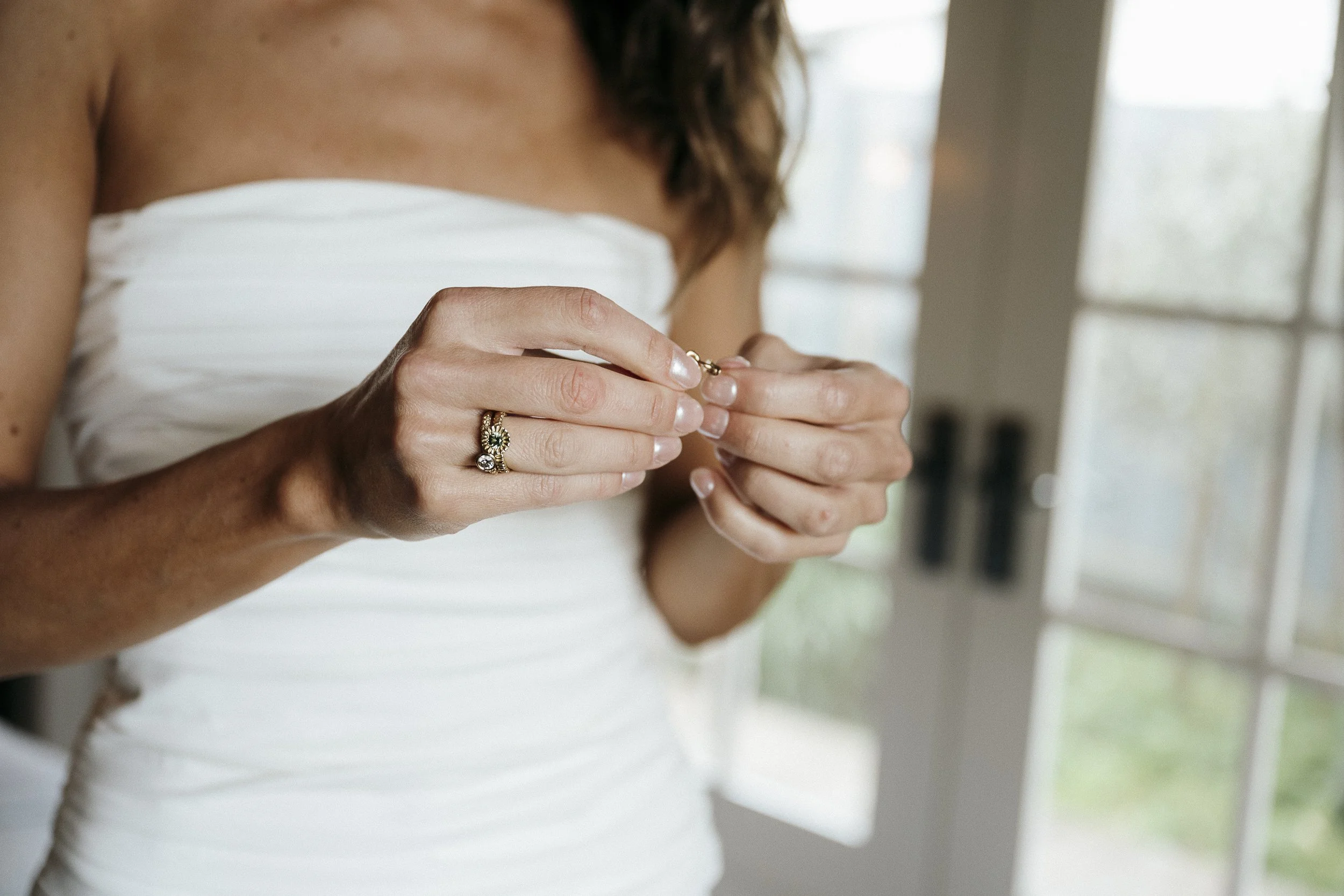 Bride standing near the window in the Miner’s Cottage in soft natural light putting on jewellery detail shot of hands.