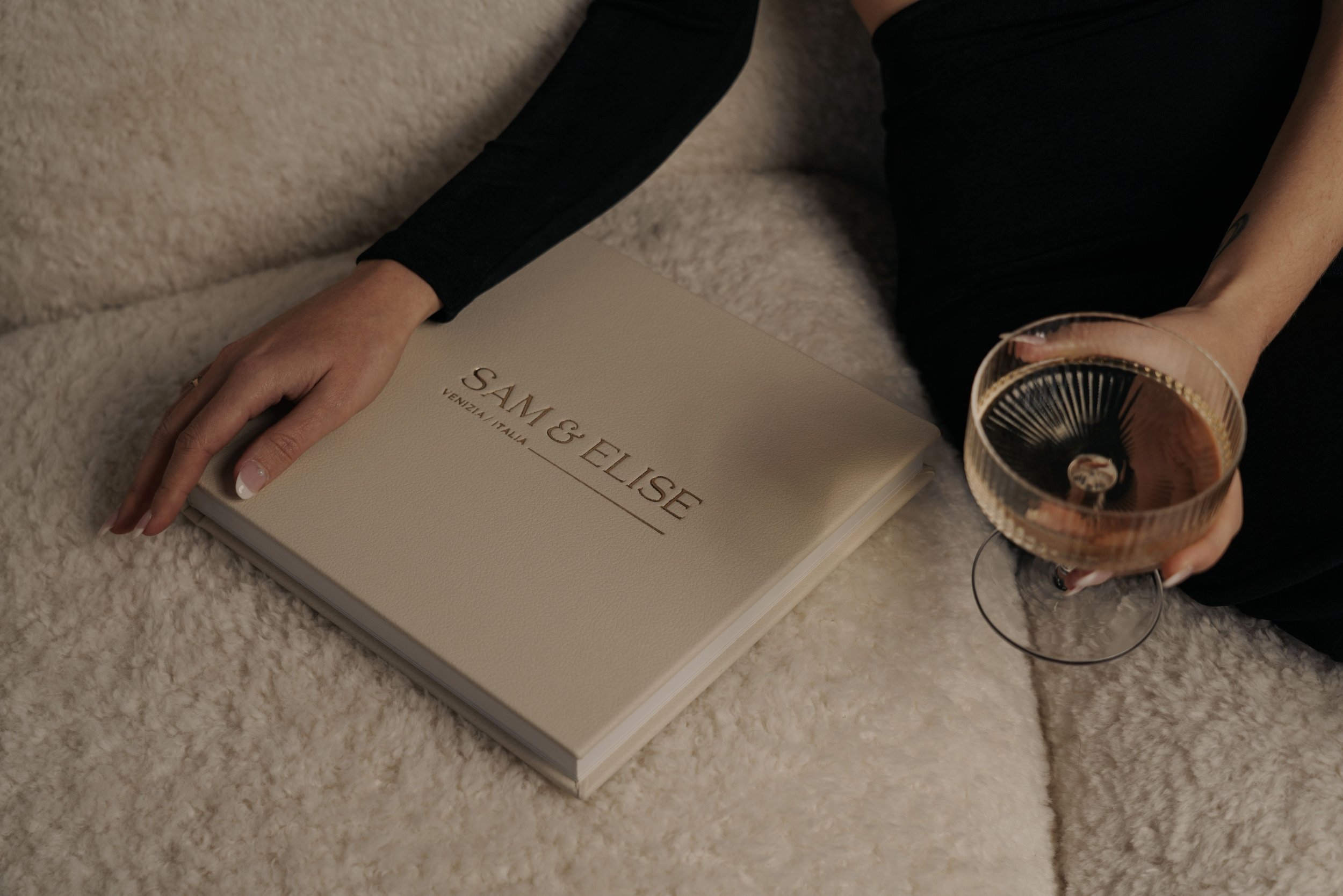 Person holding a closed beige book with "SAM & ELISE, VENEZIA, ITALIA" embossed on it, seated on a soft surface, next to a hand holding a glass of sparkling wine.