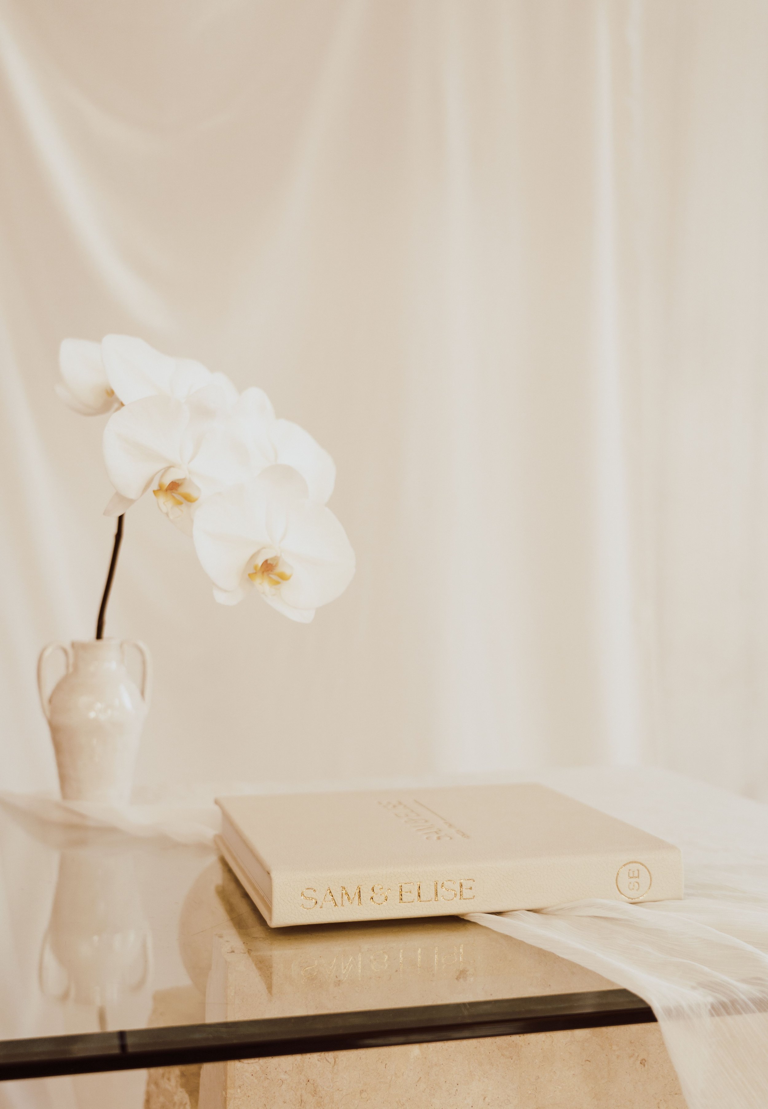 White orchids in a vase near a beige book on a glass table.