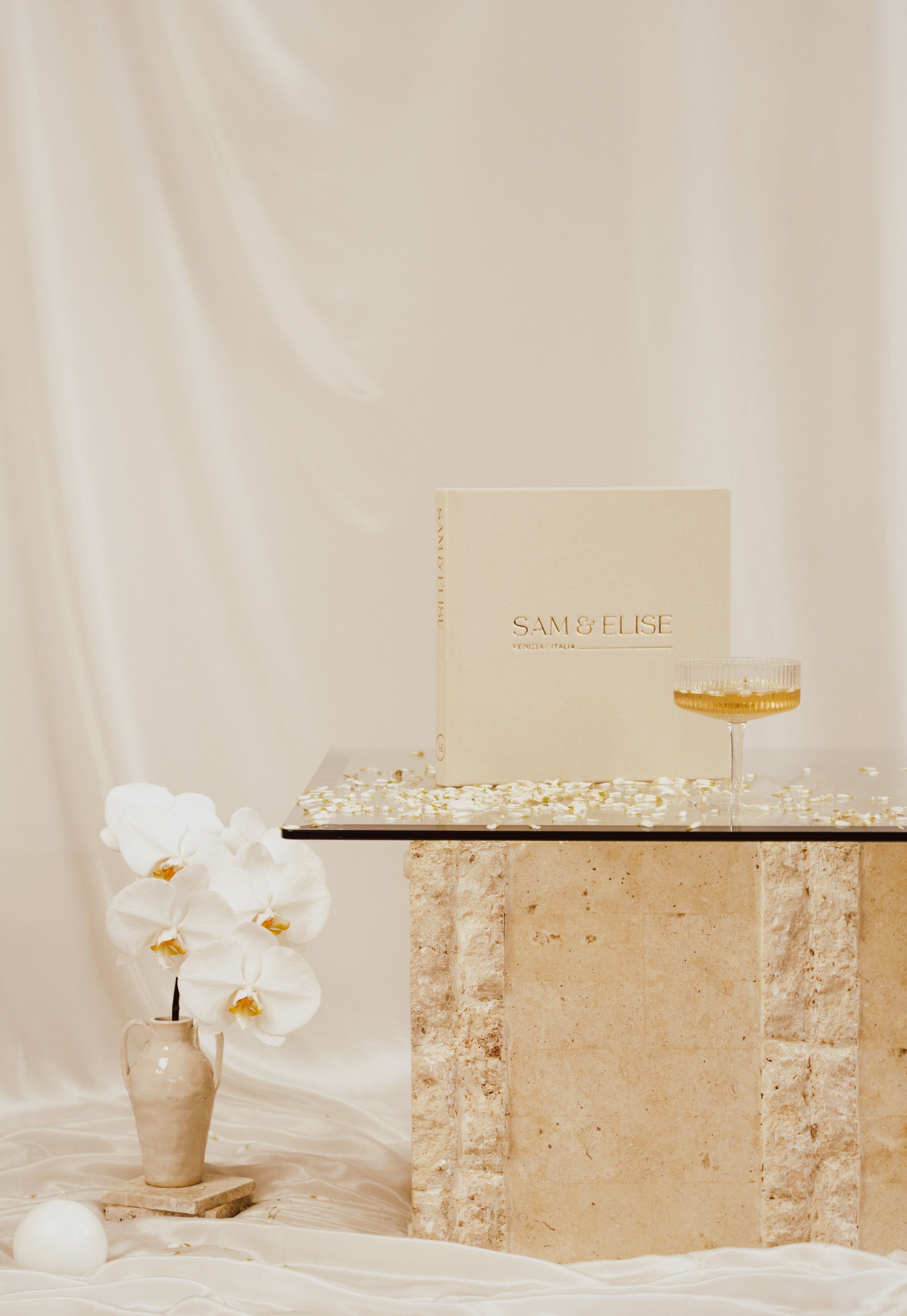 Elegant wedding album setup with title "Sam & Elise" displayed on a stone table, surrounded by scattered petals, a vase of white flowers, and a glass of champagne.
